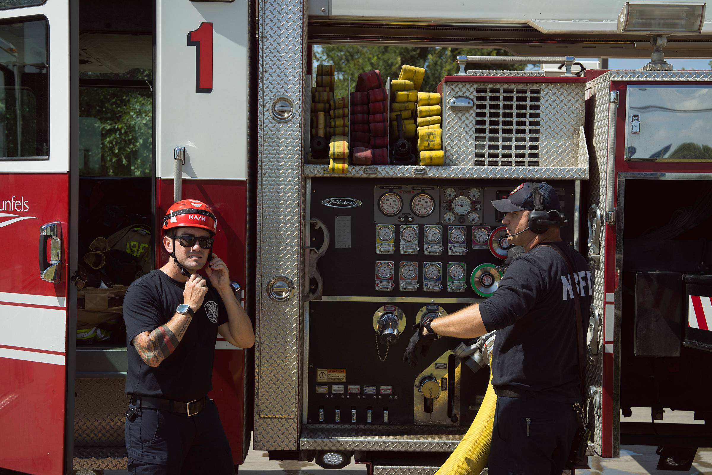 A firefighter stands outside a fire truck putting on a safety helmet. Another firefighter looks on.