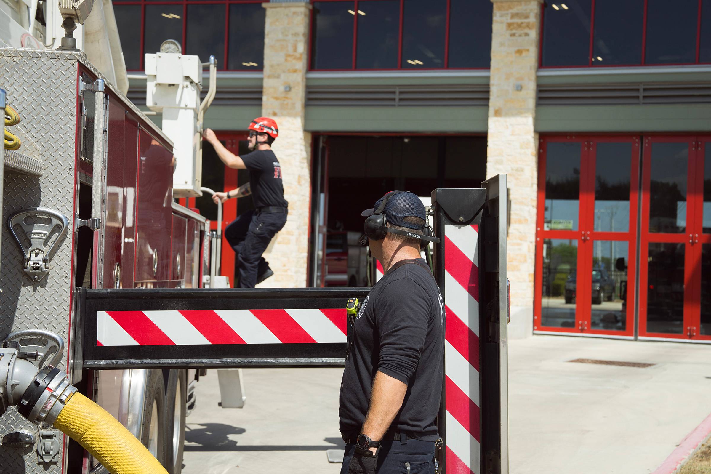 A firefighter wearing a safety helmet climbs onto the truck. Another firefighter in the foreground looks on.