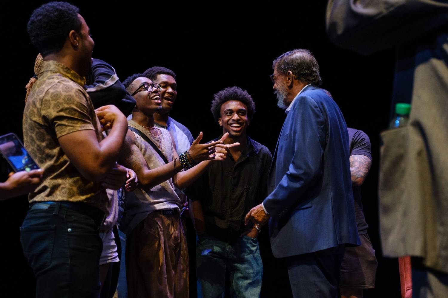 A group of people gather on a dark theater stage, smiling and talking together after a performance. An older man in a blue suit faces several younger men at center stage.