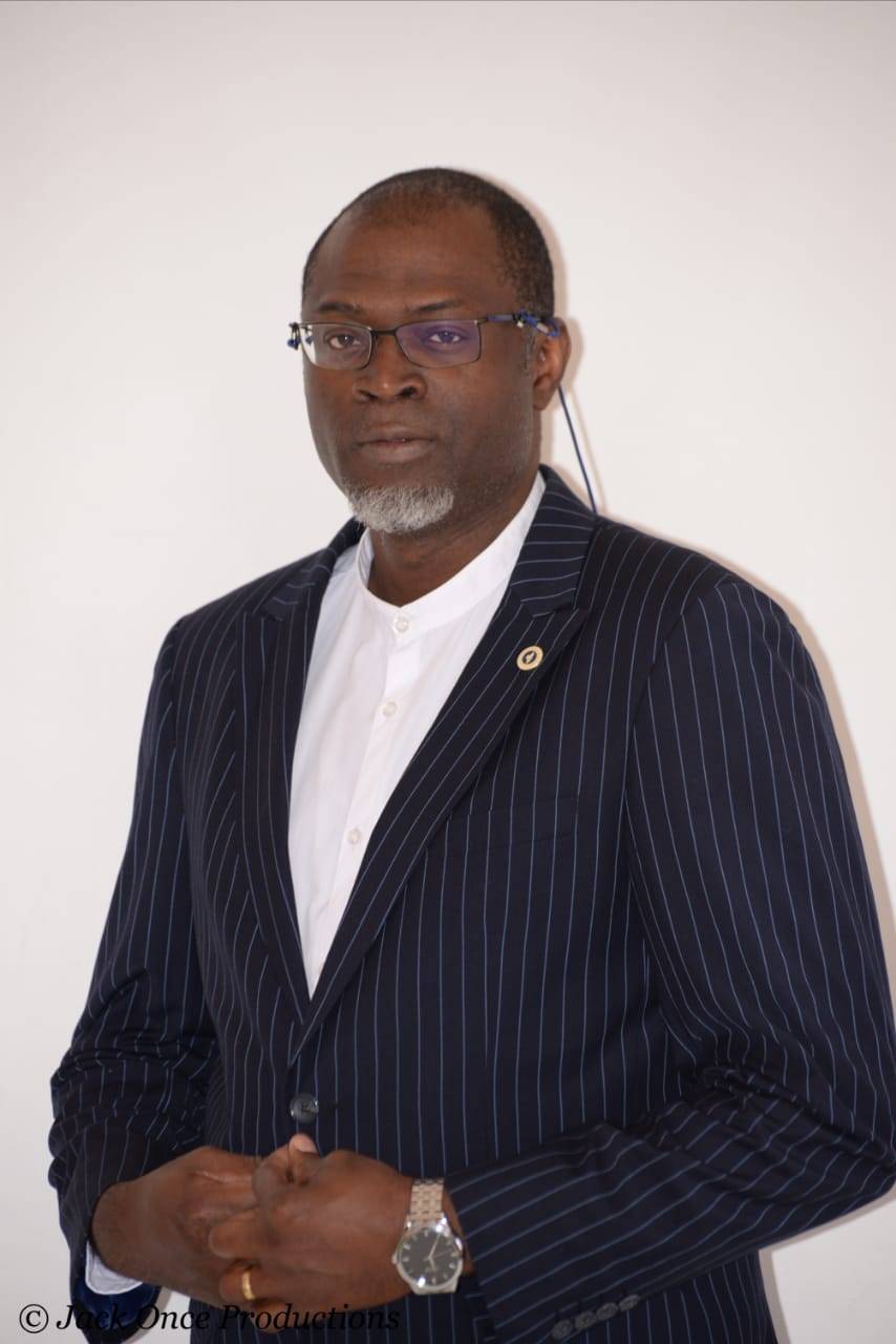 Professional portrait of a person wearing glasses, a white shirt, and a navy pinstripe blazer, standing against a plain light-colored background.