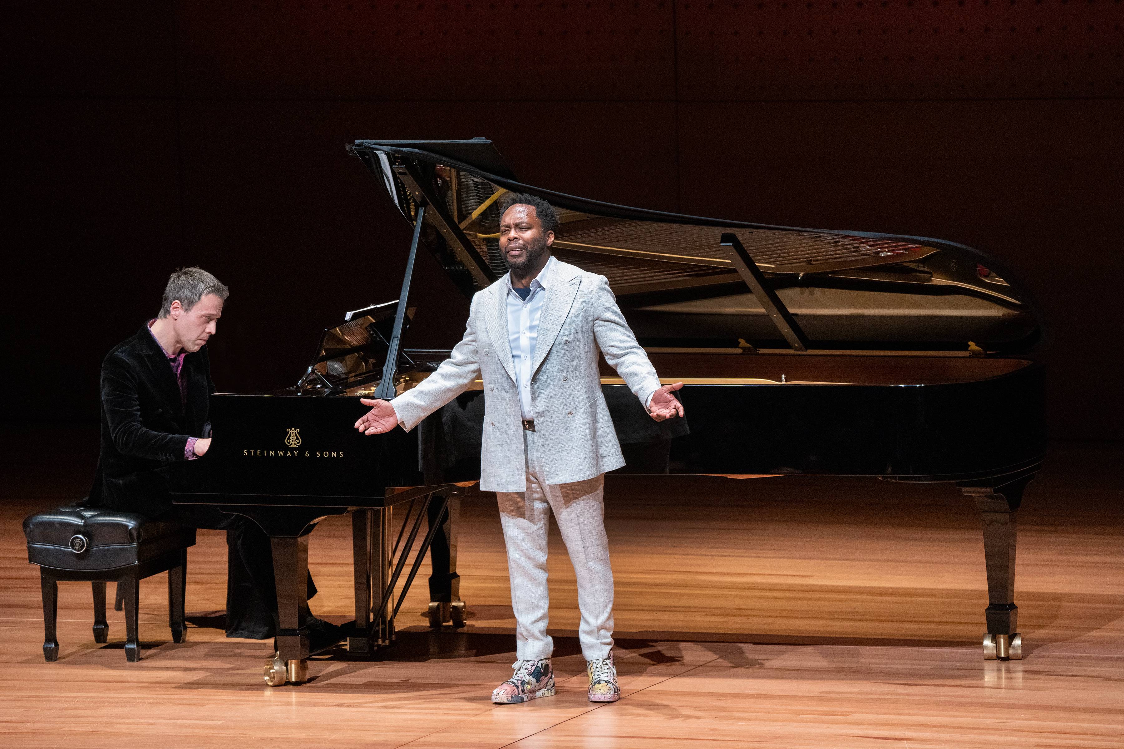man in light colored suit sings on stage in front of large piano 