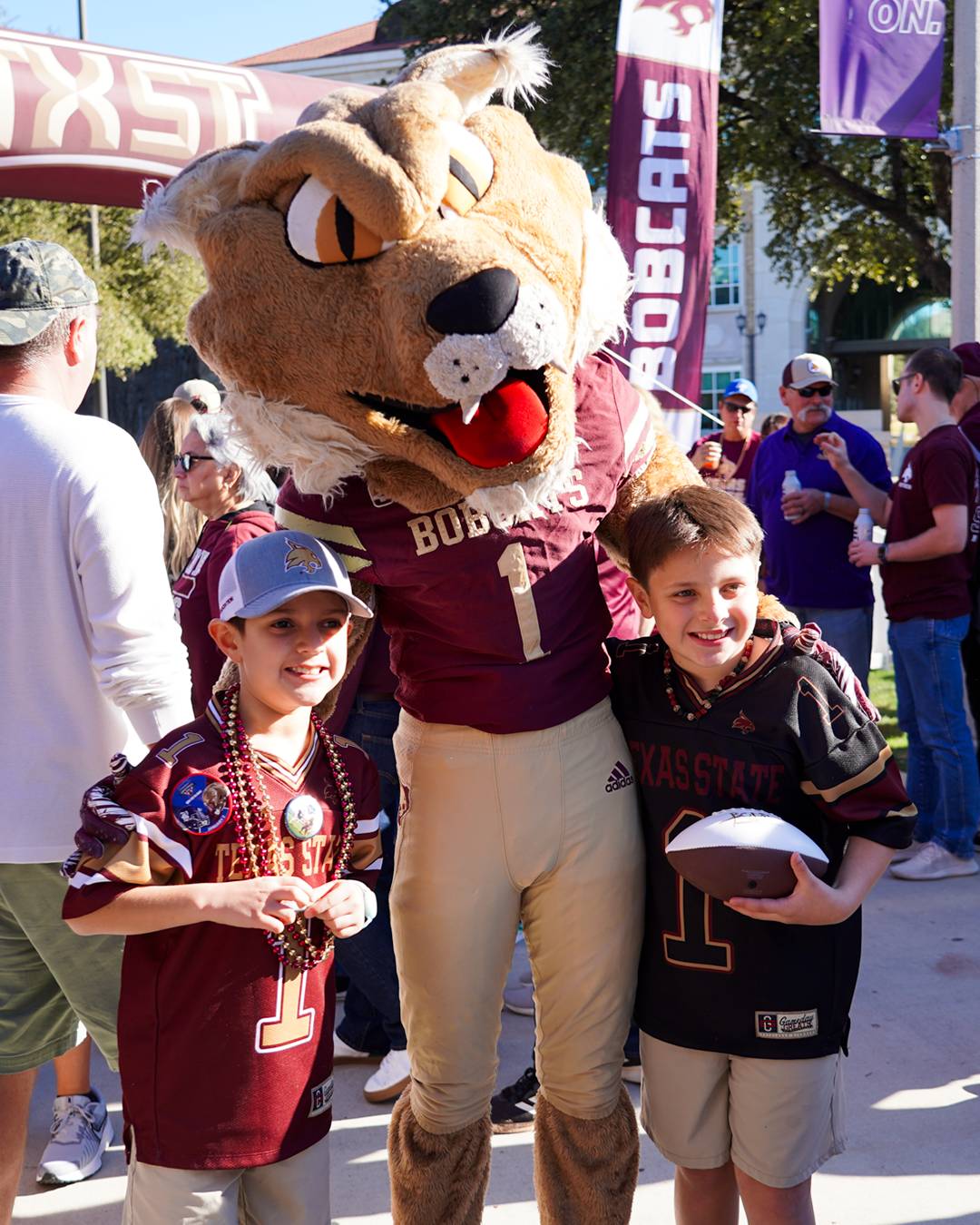 Two young Bobcat fans posing for a photo with Boko the Bobcat mascot at a Texas State football event
