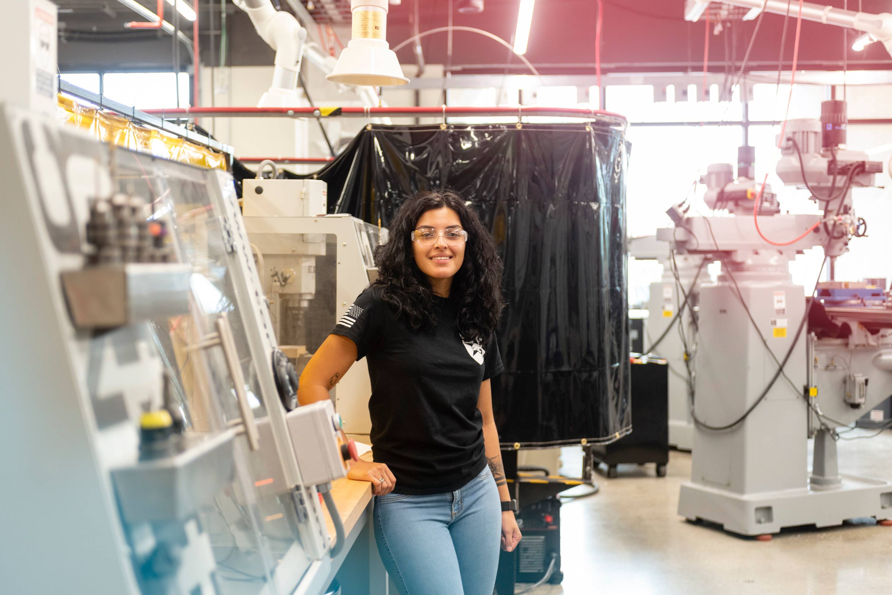 A woman wearing glasses and a white lab coat over a plaid shirt smiles at the camera while standing in a laboratory, with scientific instruments and equipment visible in the background.