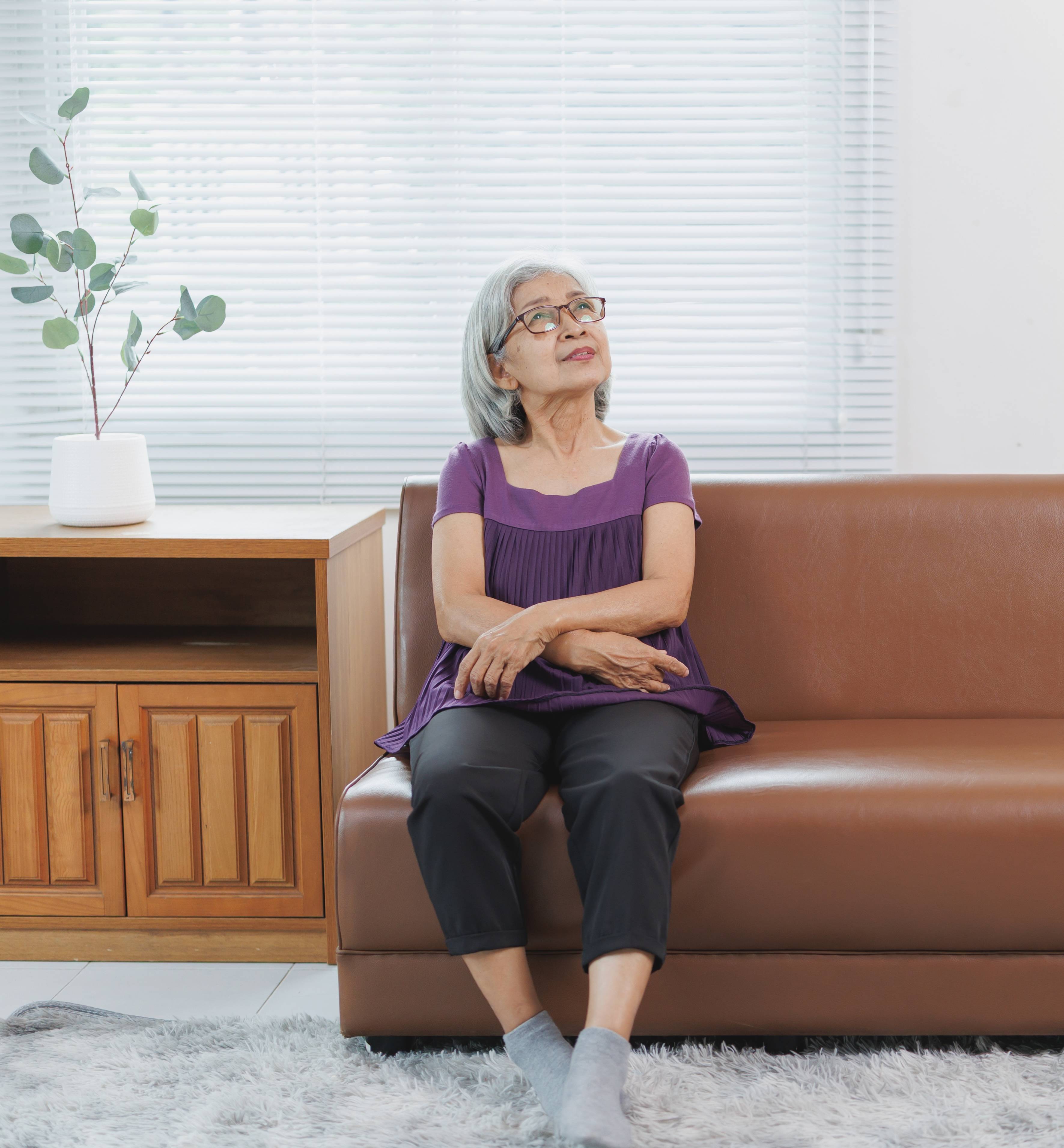 An elderly woman in a purple blouse sites on a couch and looks up hopefully.