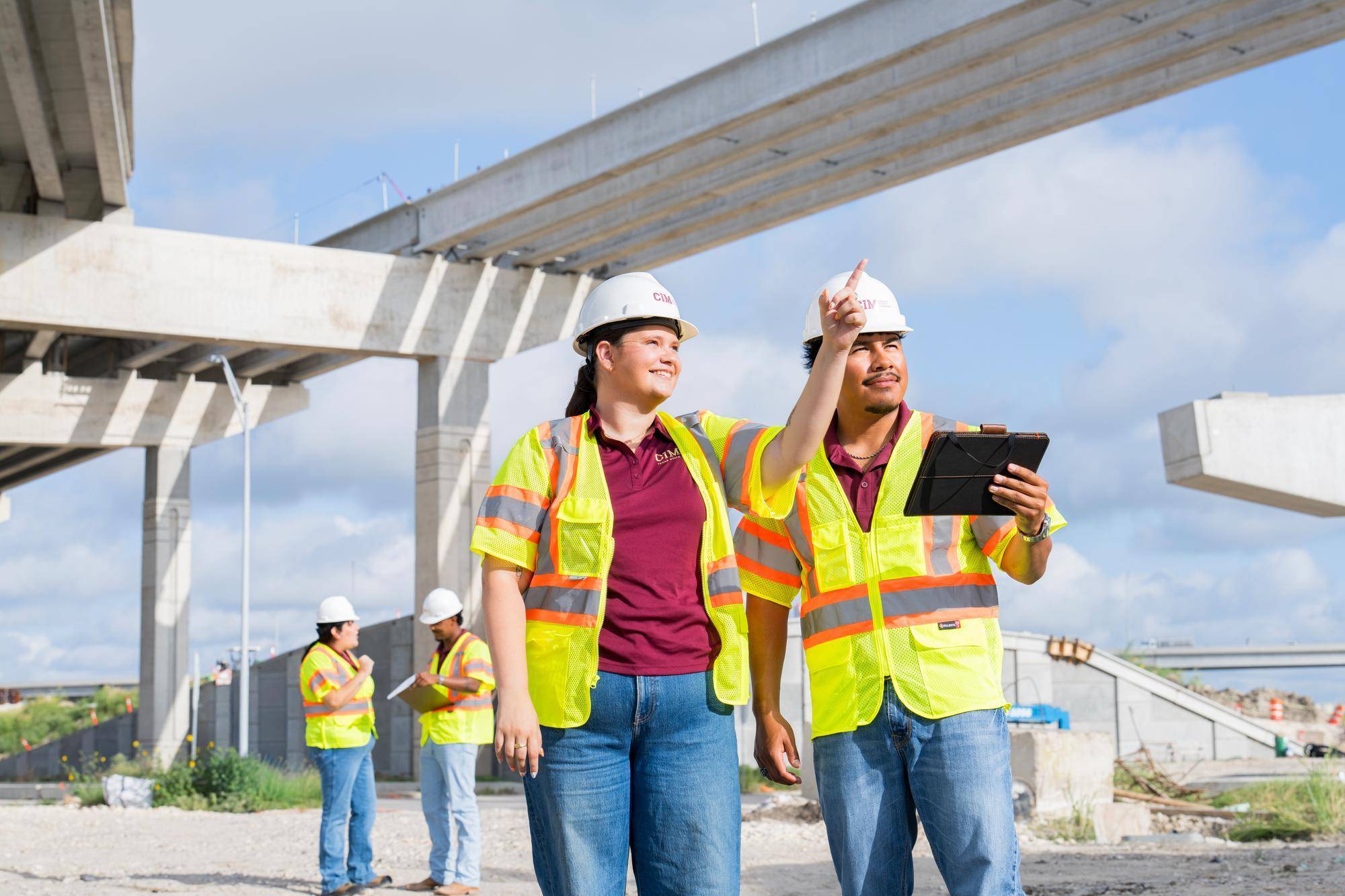 four people in yellow safety vests on a construction site under bridge.