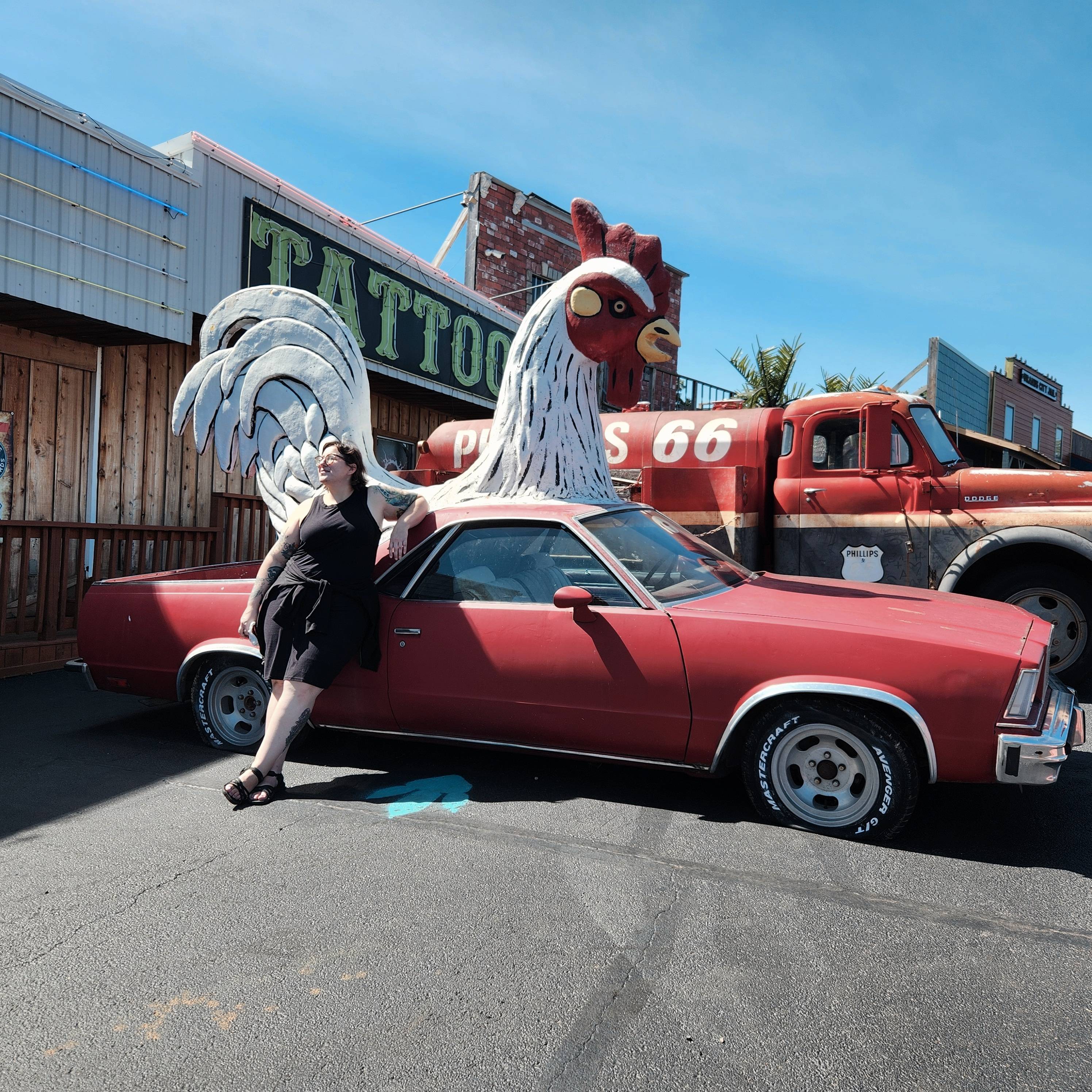 picture of Ashley with a vintage car that has a giant rooster on top and a tattoo shop behind