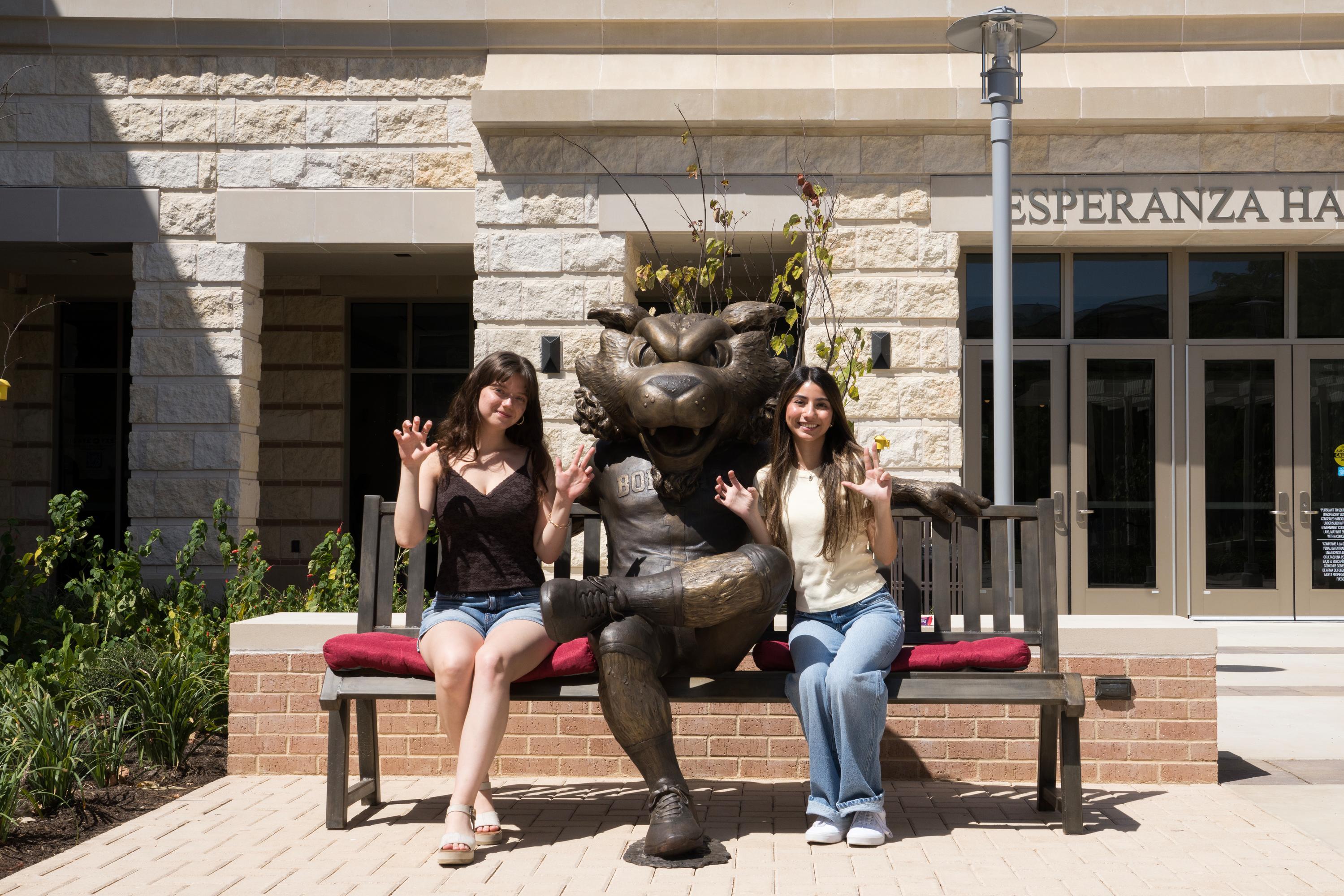 two women sit on either size of a large bronze bobcat statue that is positioned on a bronze bench