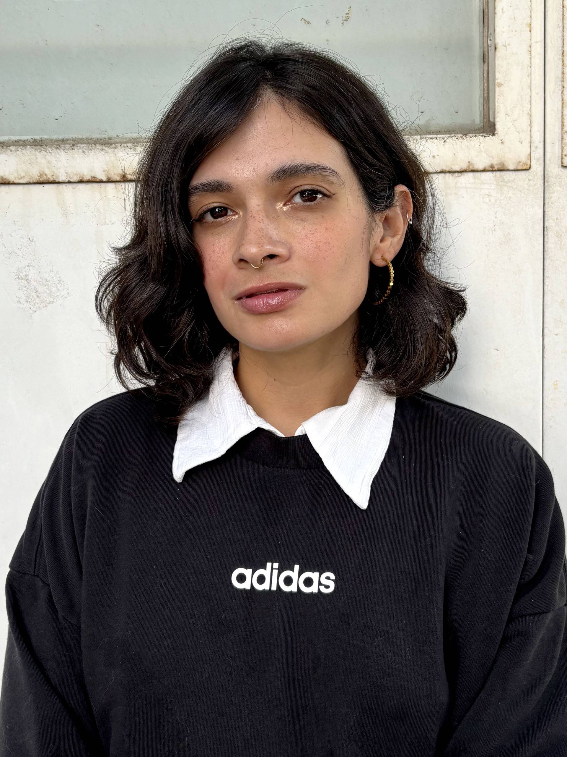 A dark haired woman from the waist up standing in front of a white wall.