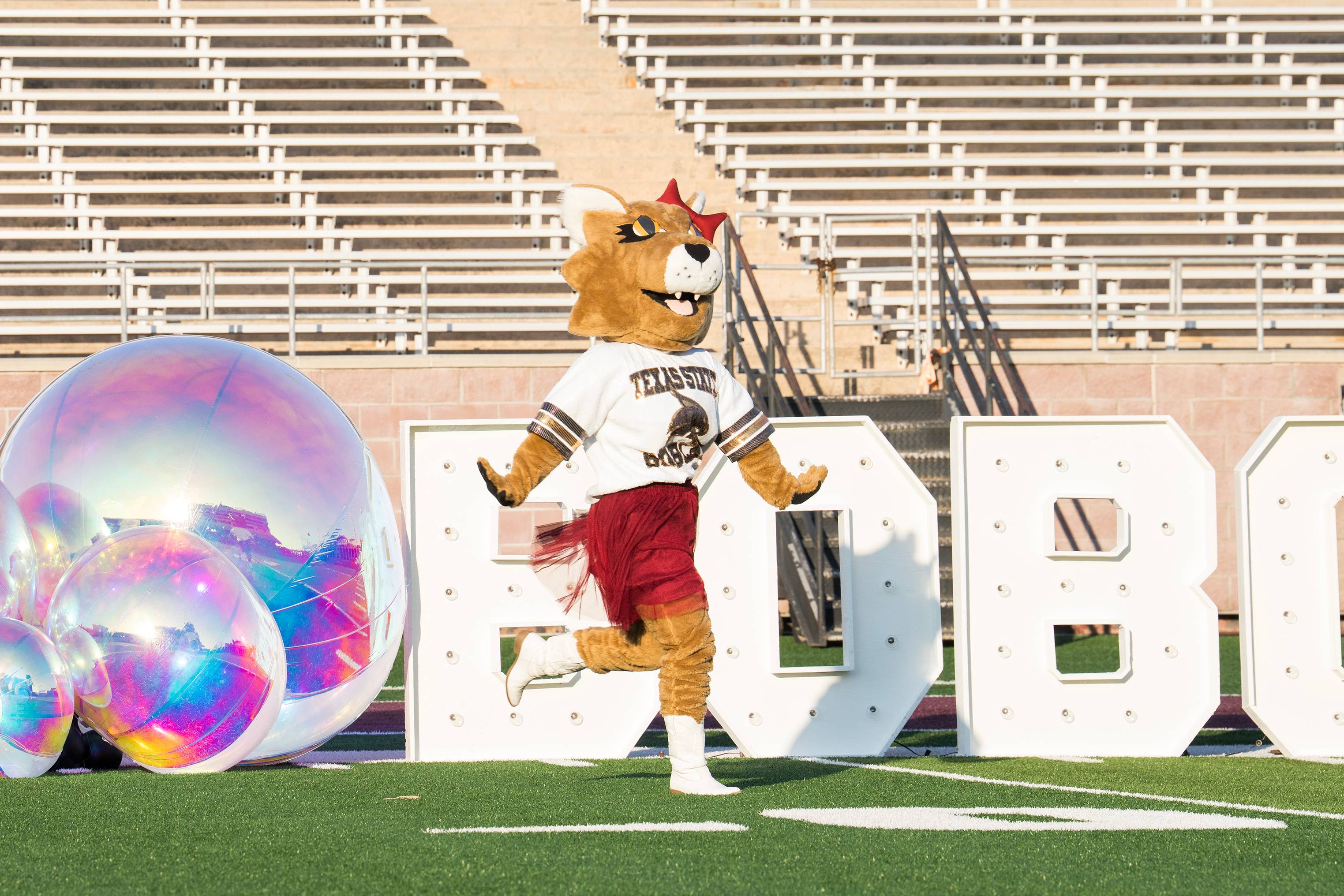 female bobcat mascot runs onto football field in front of large blow rainbow bubbles