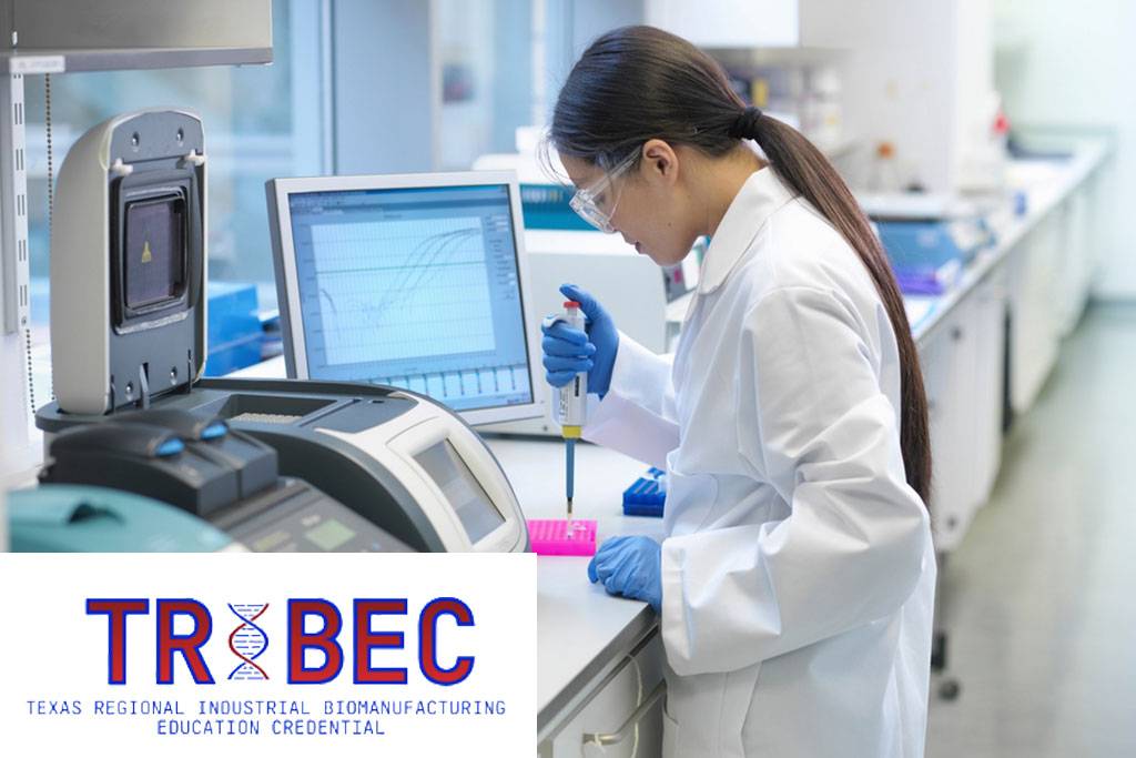 A woman wearing glasses and a white lab coat over a plaid shirt smiles at the camera while standing in a laboratory, with scientific instruments and equipment visible in the background."

If you'd like a shorter version for tighter character limits: "A smiling researcher in a lab coat standing in a laboratory with equipment behind him.