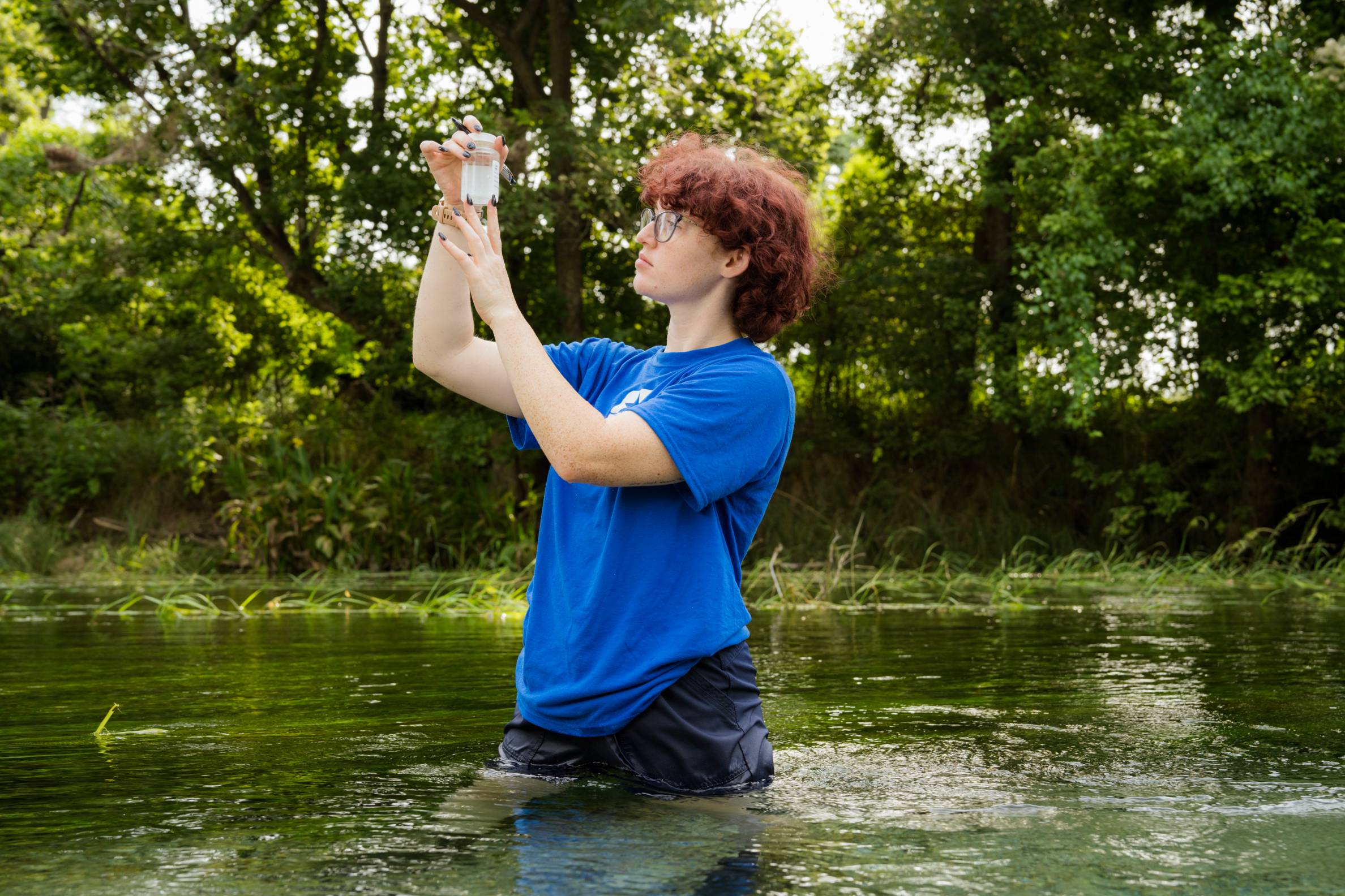 person wearing a blue shirt standing in the river holding up a container looking at water quality 
