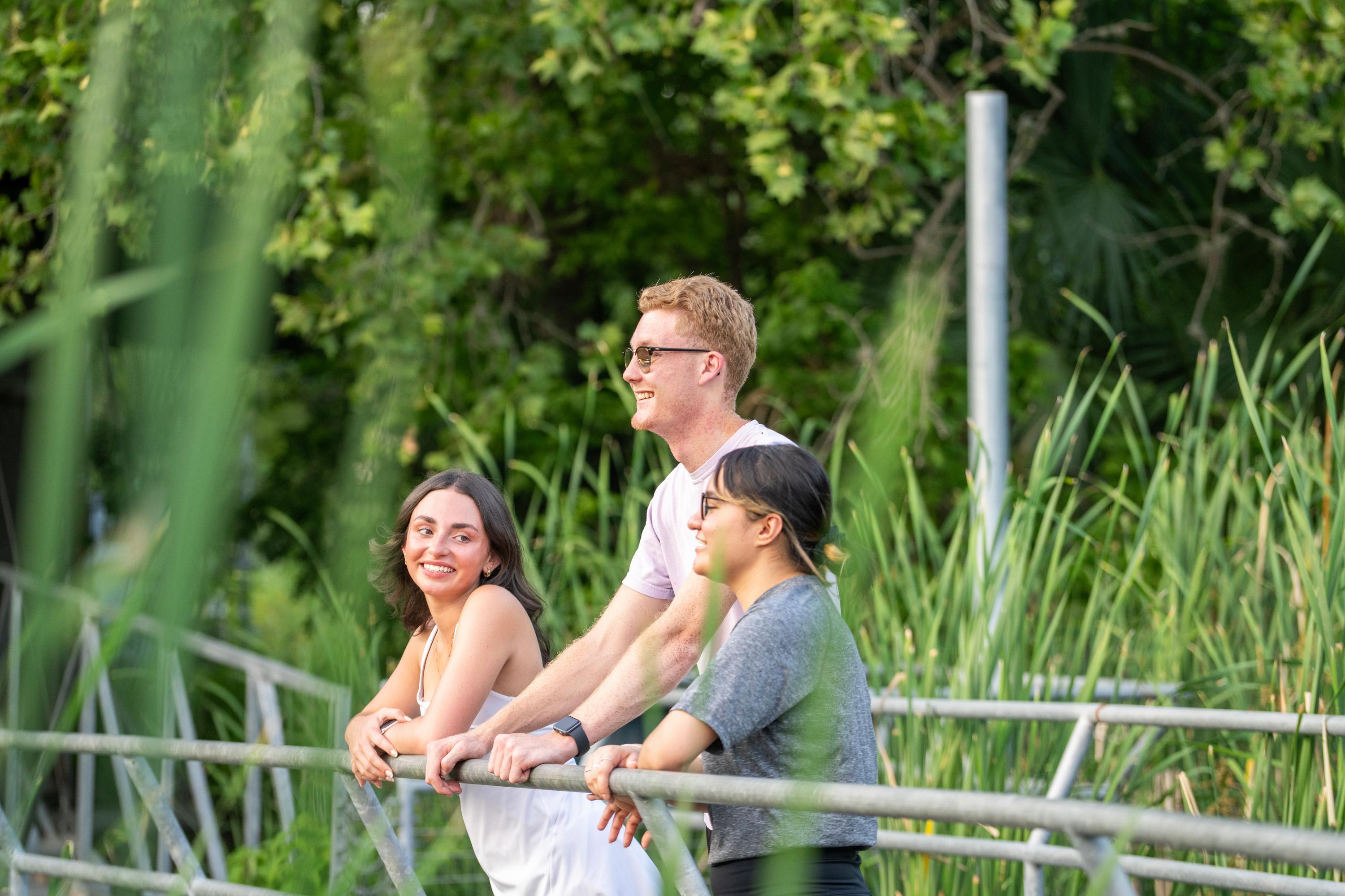 three students leaning on metal railing while surrounded by tall grass