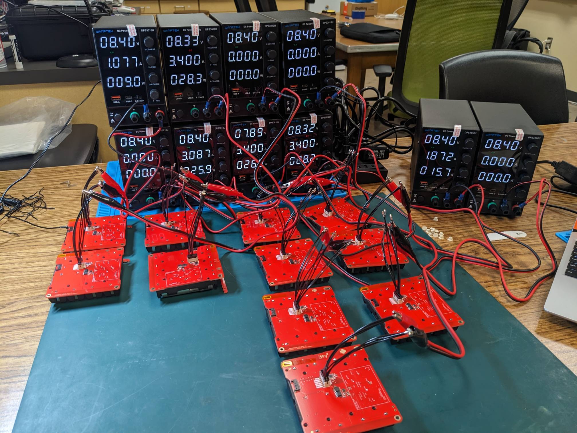 Multiple red circuit boards connected by wires to digital power supplies on a laboratory workbench.