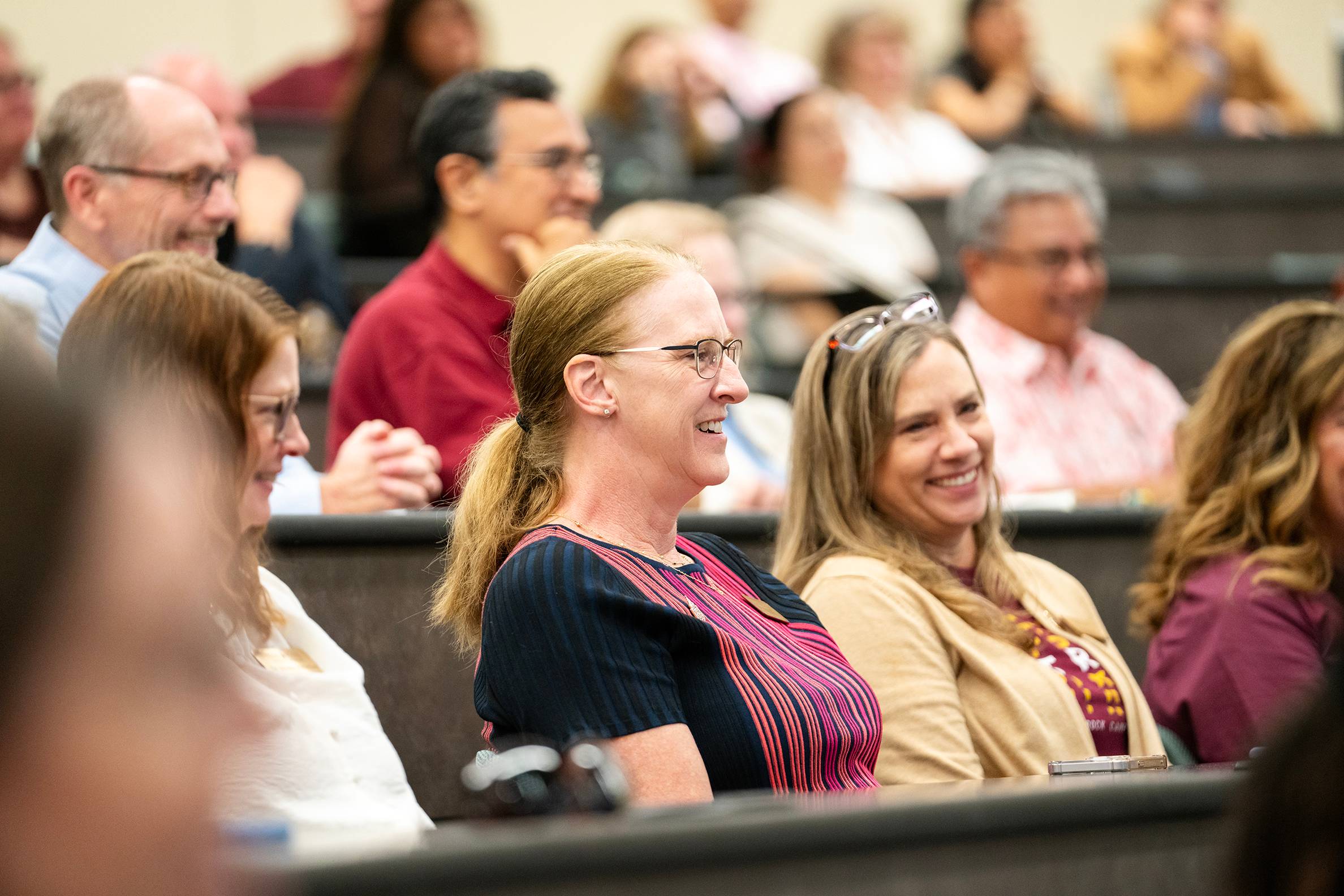 staff members sitting in auditorium laughing while listening to presentation