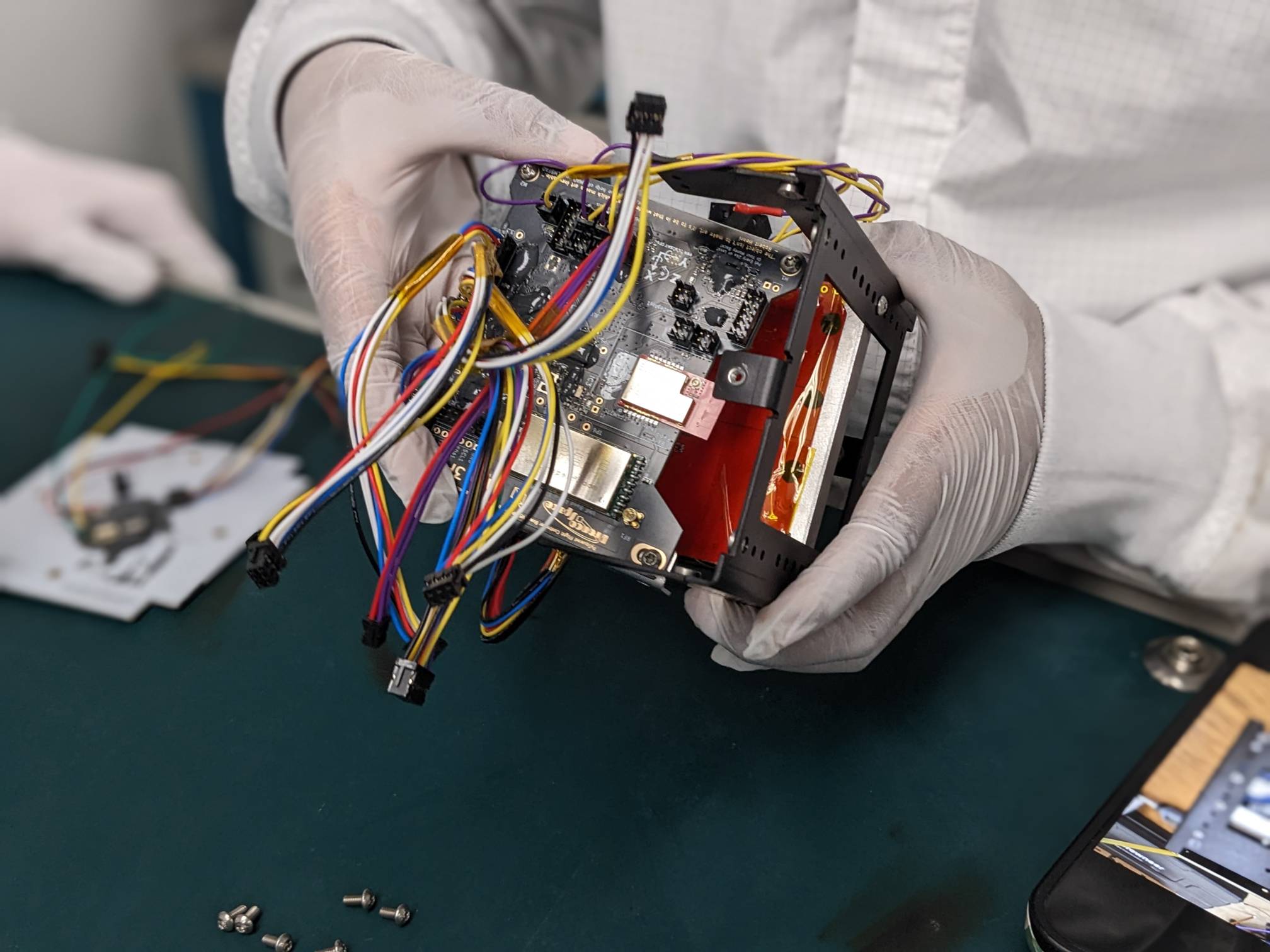 Gloved hands hold an electronic device with exposed circuit boards and multicolored wiring.