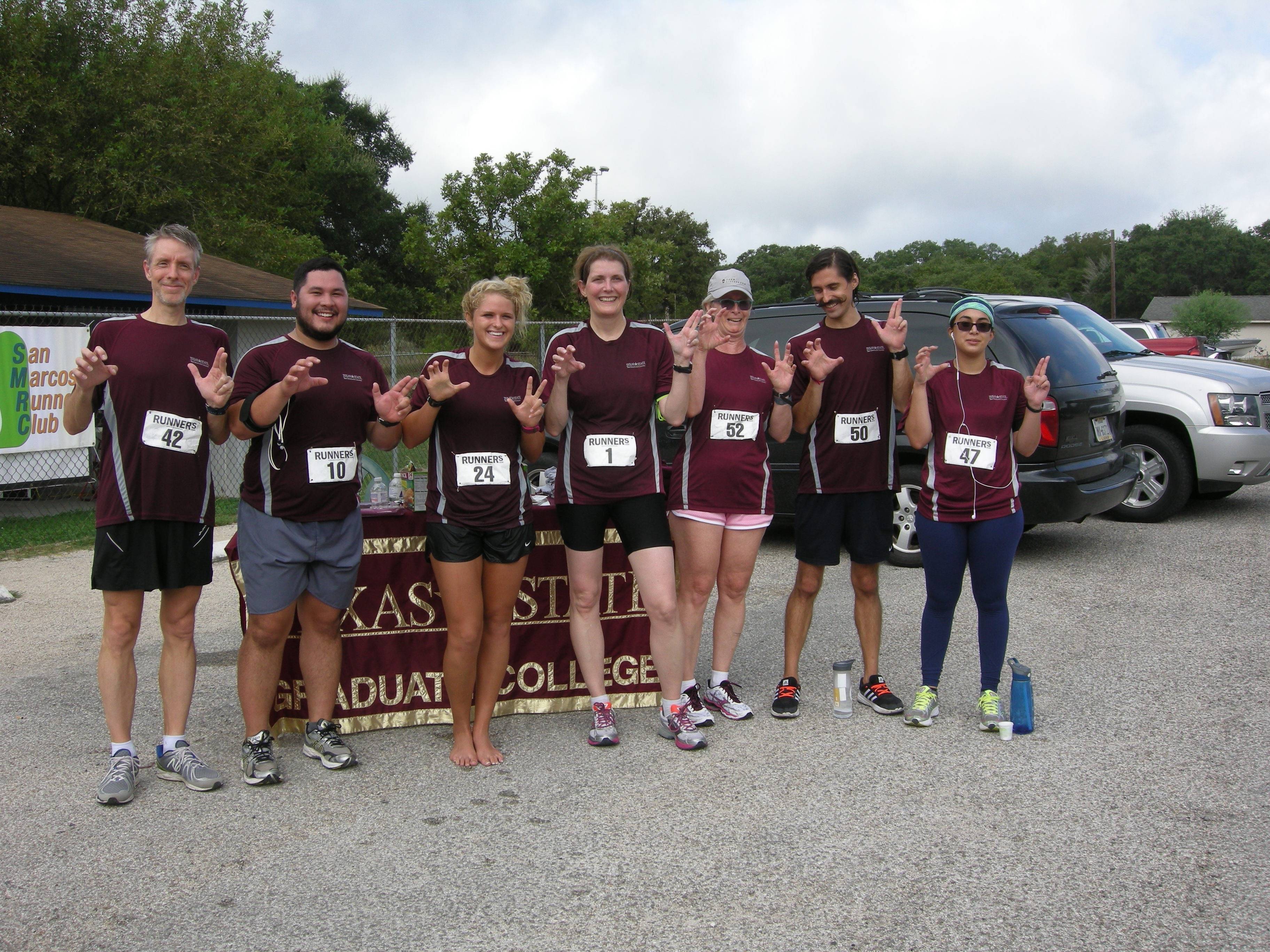 seven people in maroon shirts and running attire holding up txst hand signs