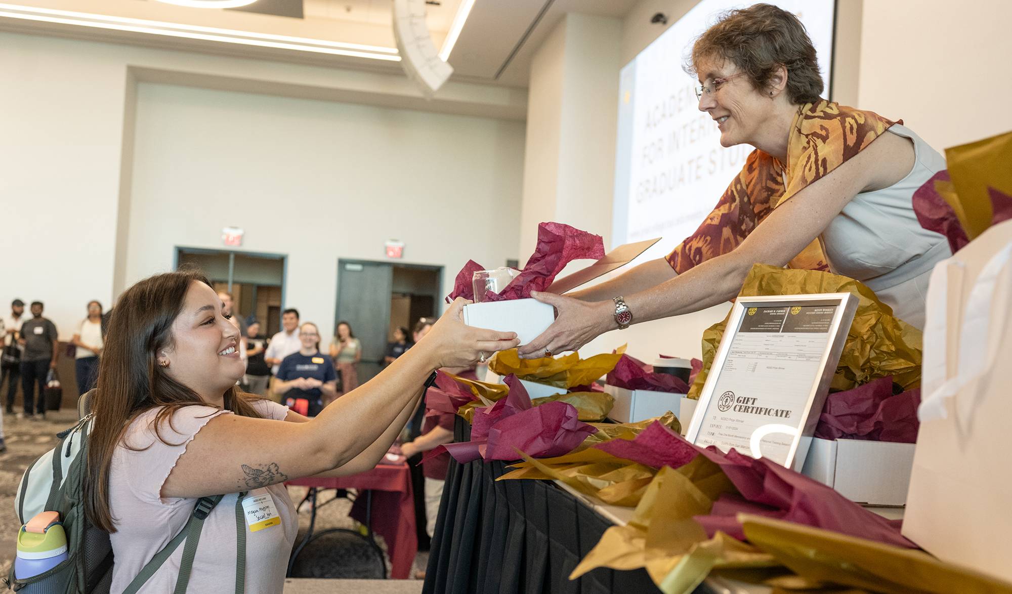 A woman hands a gift box to a student during an event as attendees look on.