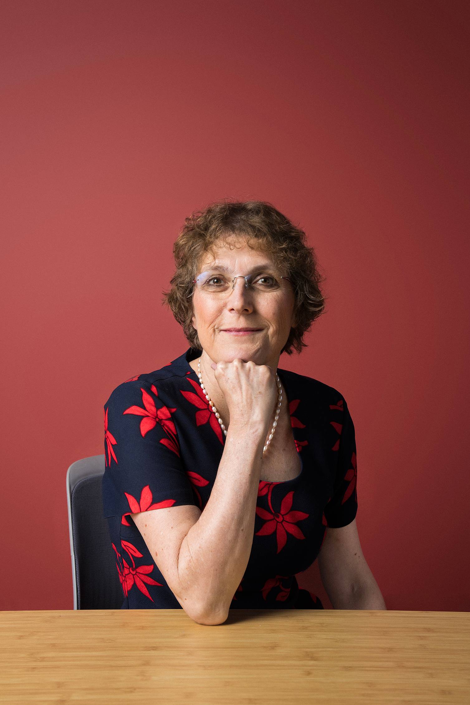 Portrait of a woman wearing glasses and a black dress with red flowers, resting her chin on her hand against a red background.