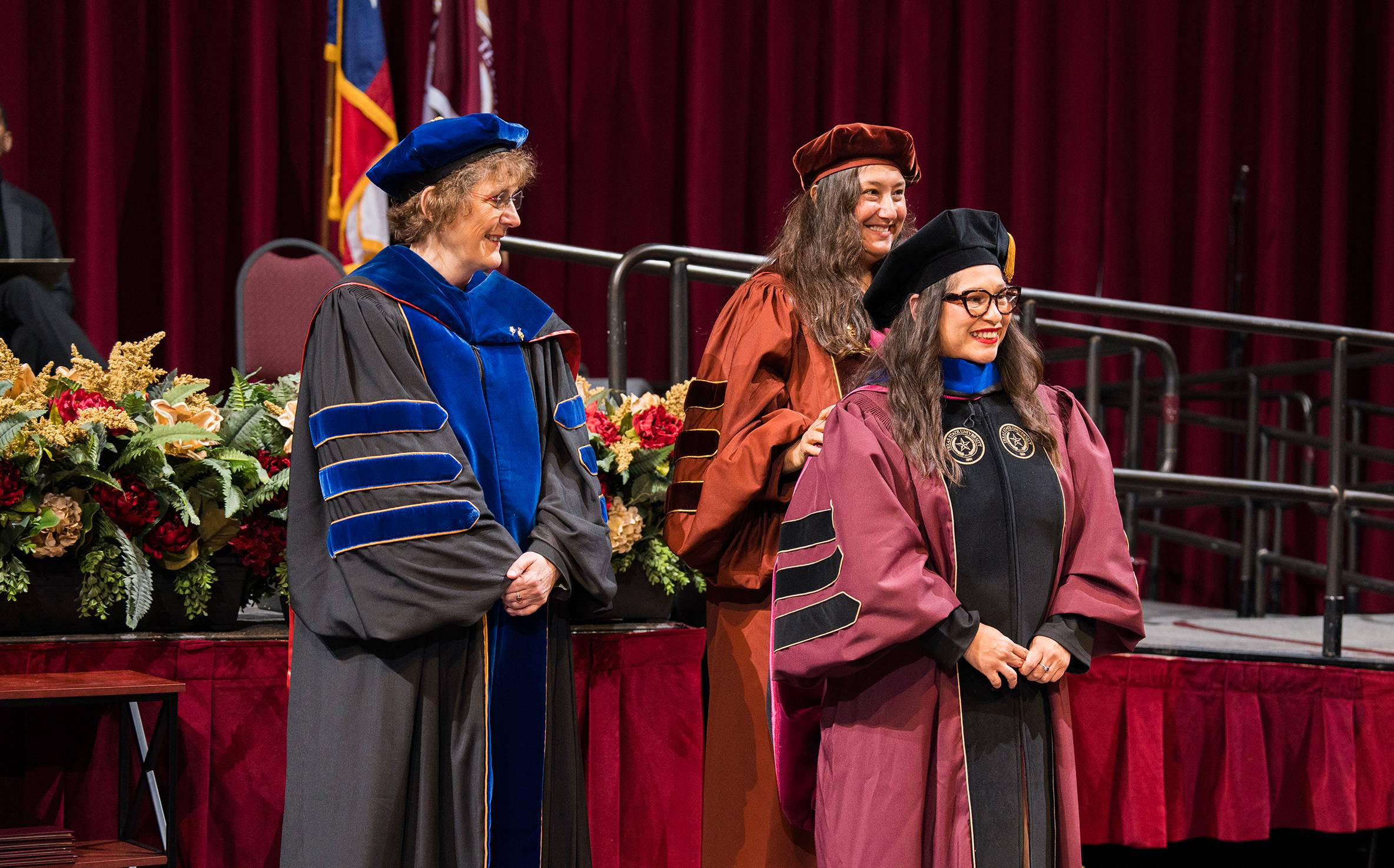 Three women in academic regalia smile together onstage during a commencement ceremony.