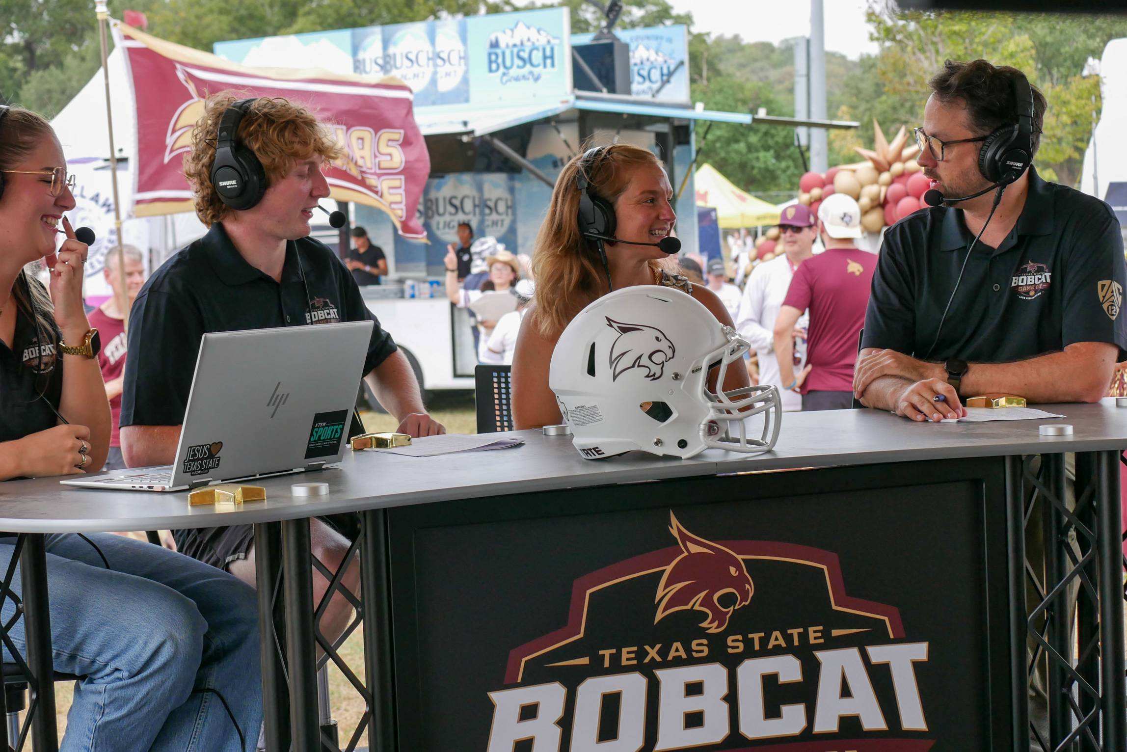 Bobcat Game Day hosts wearing headsets talk at an outdoor broadcast desk with a Texas State football helmet.