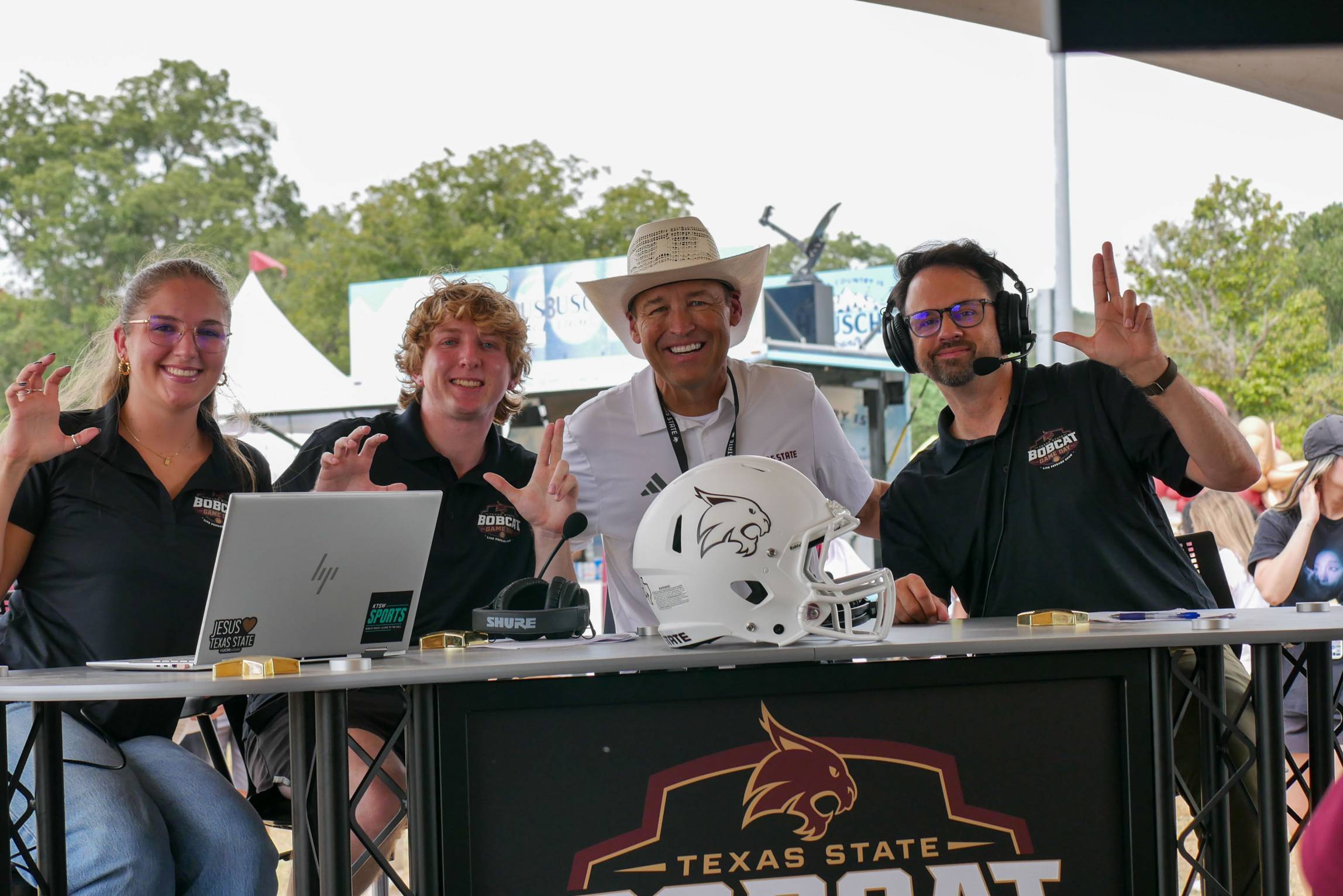 Bobcat Game Day hosts pose together behind the broadcast desk and make the Texas State hand sign.