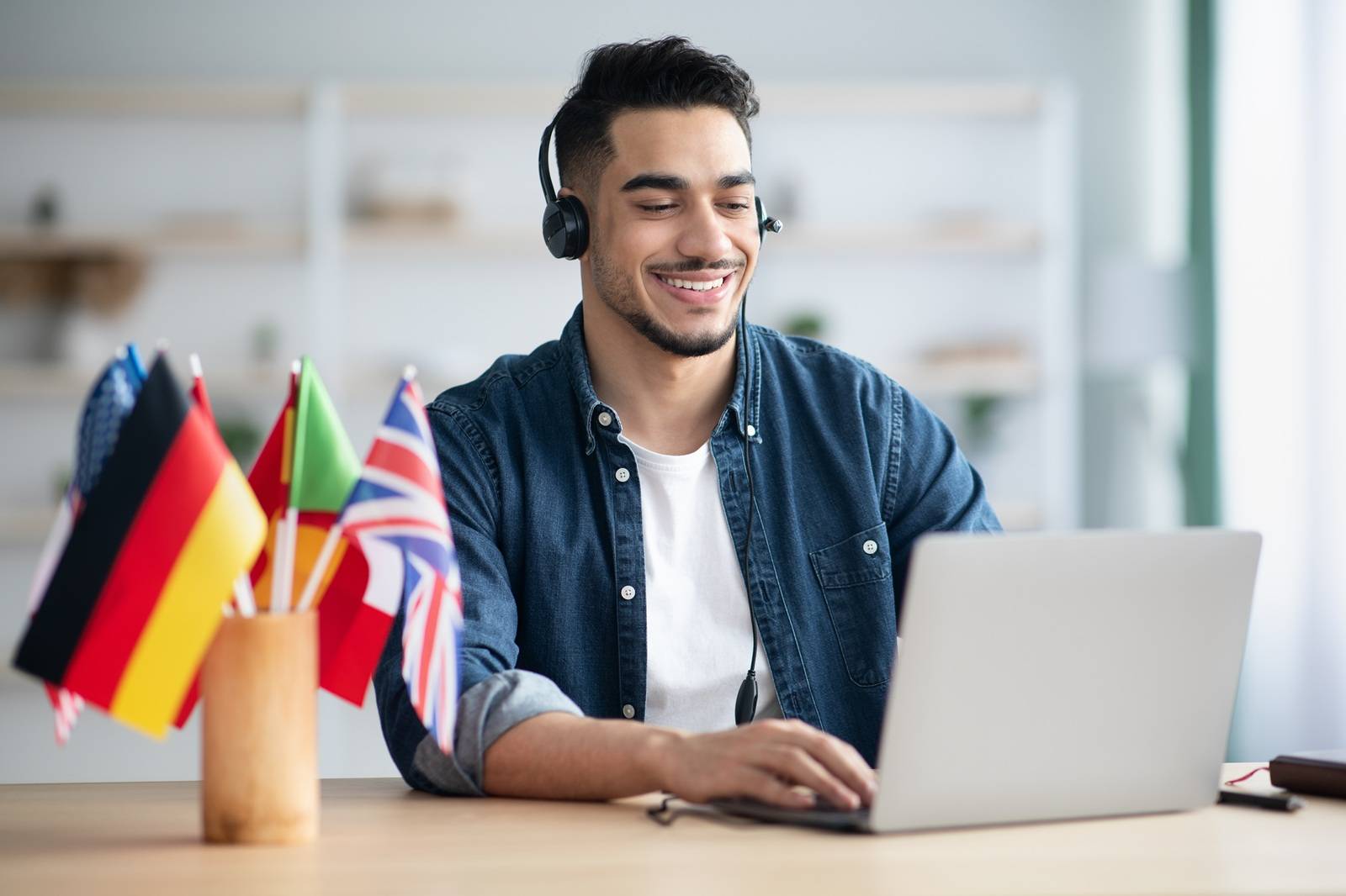 man wearing headphones, smiling, sitting in front of laptop, glass on table with various flags