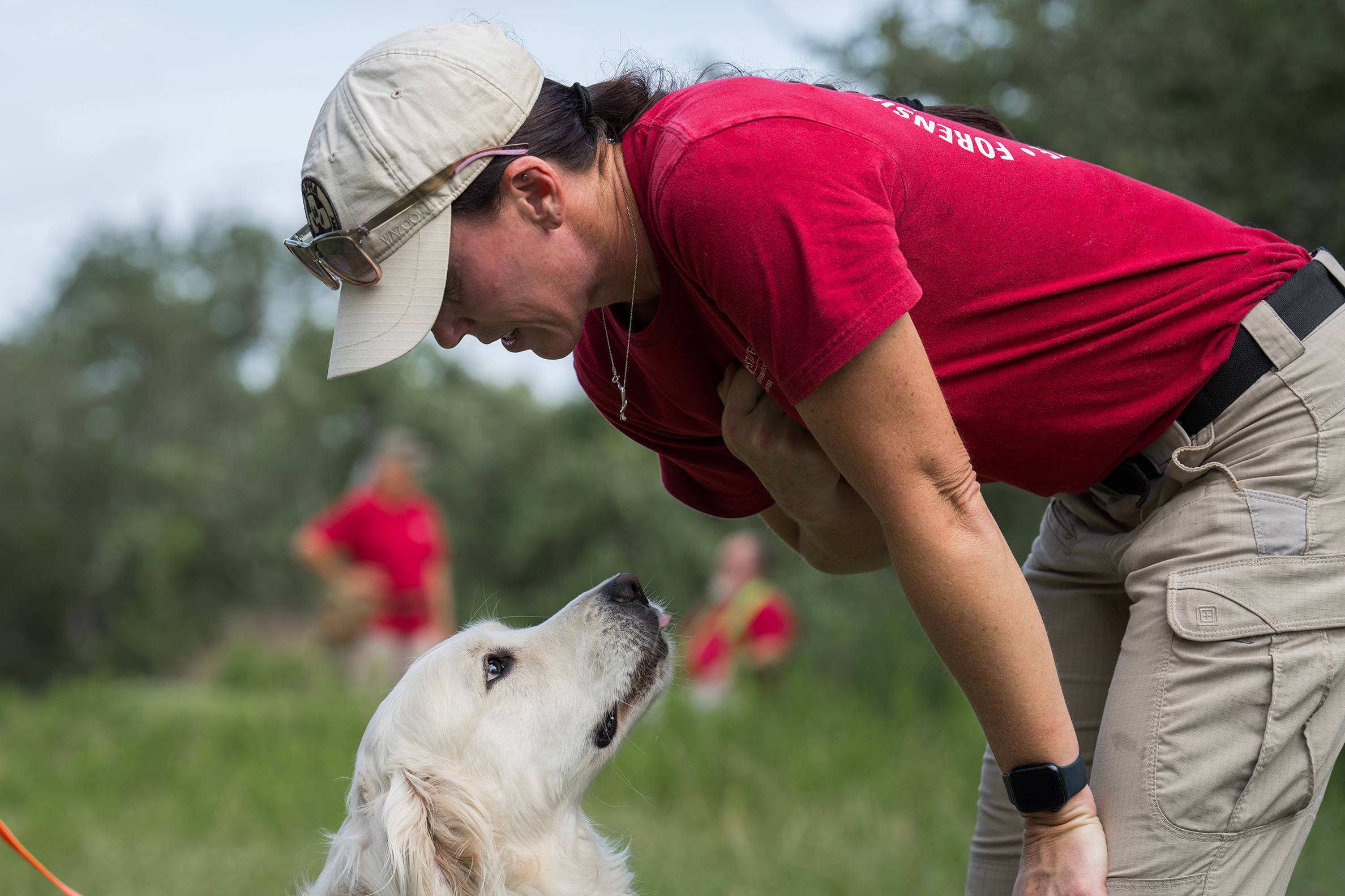 woman in baseball hat and red shirt bends down to look at white dog licking its lips