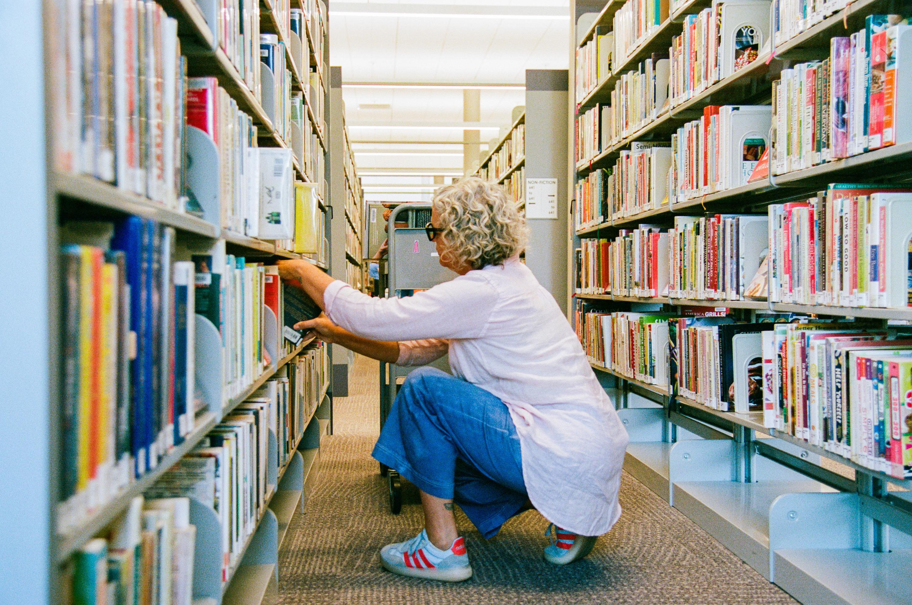 woman bending down in between two rows of books at a library. she is restocking the shelves with books. there is a cart of books besides her