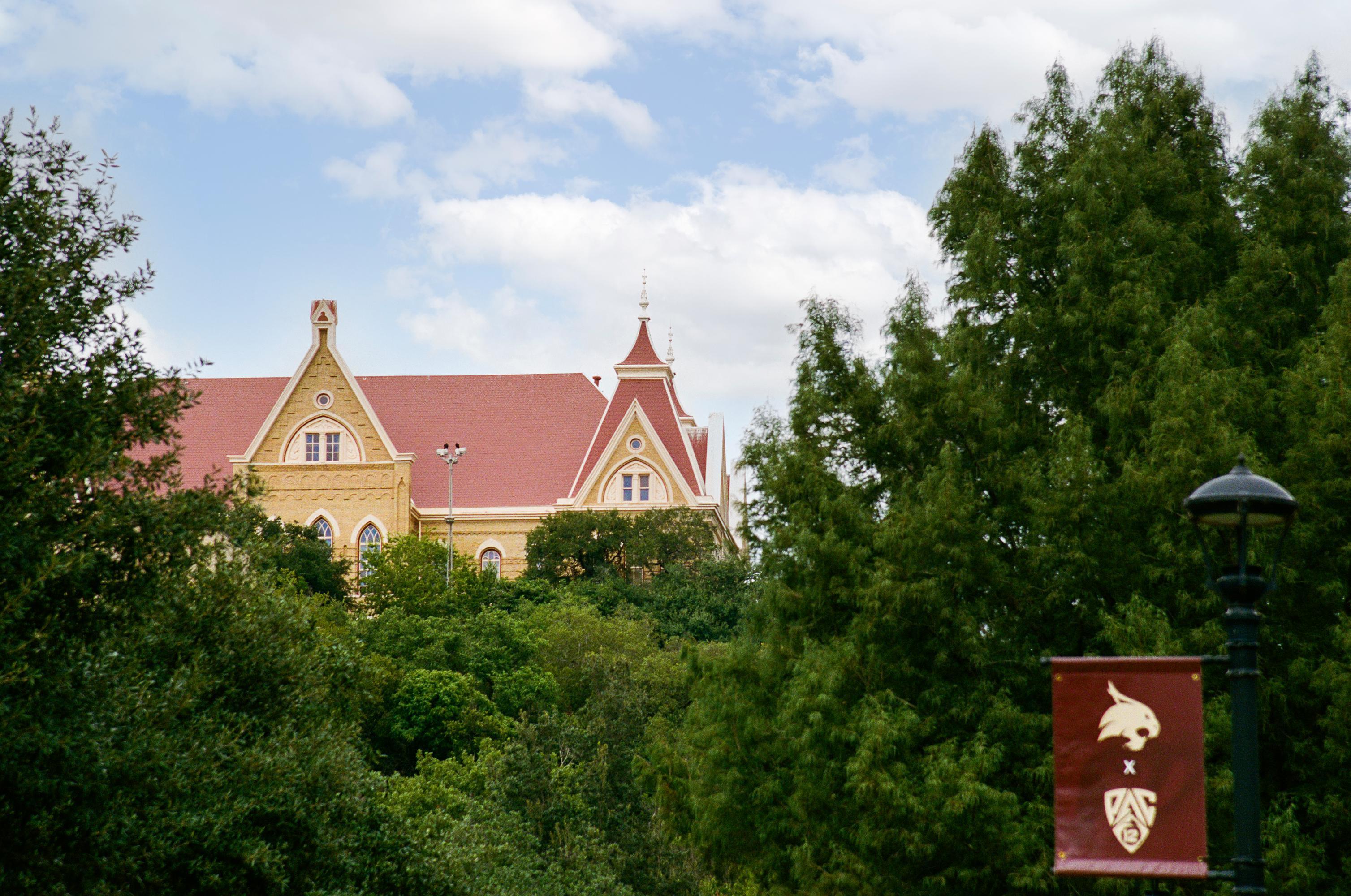 the red roof of Old Main peaks behind green trees. a blue sky with clouds is shown behind it. 