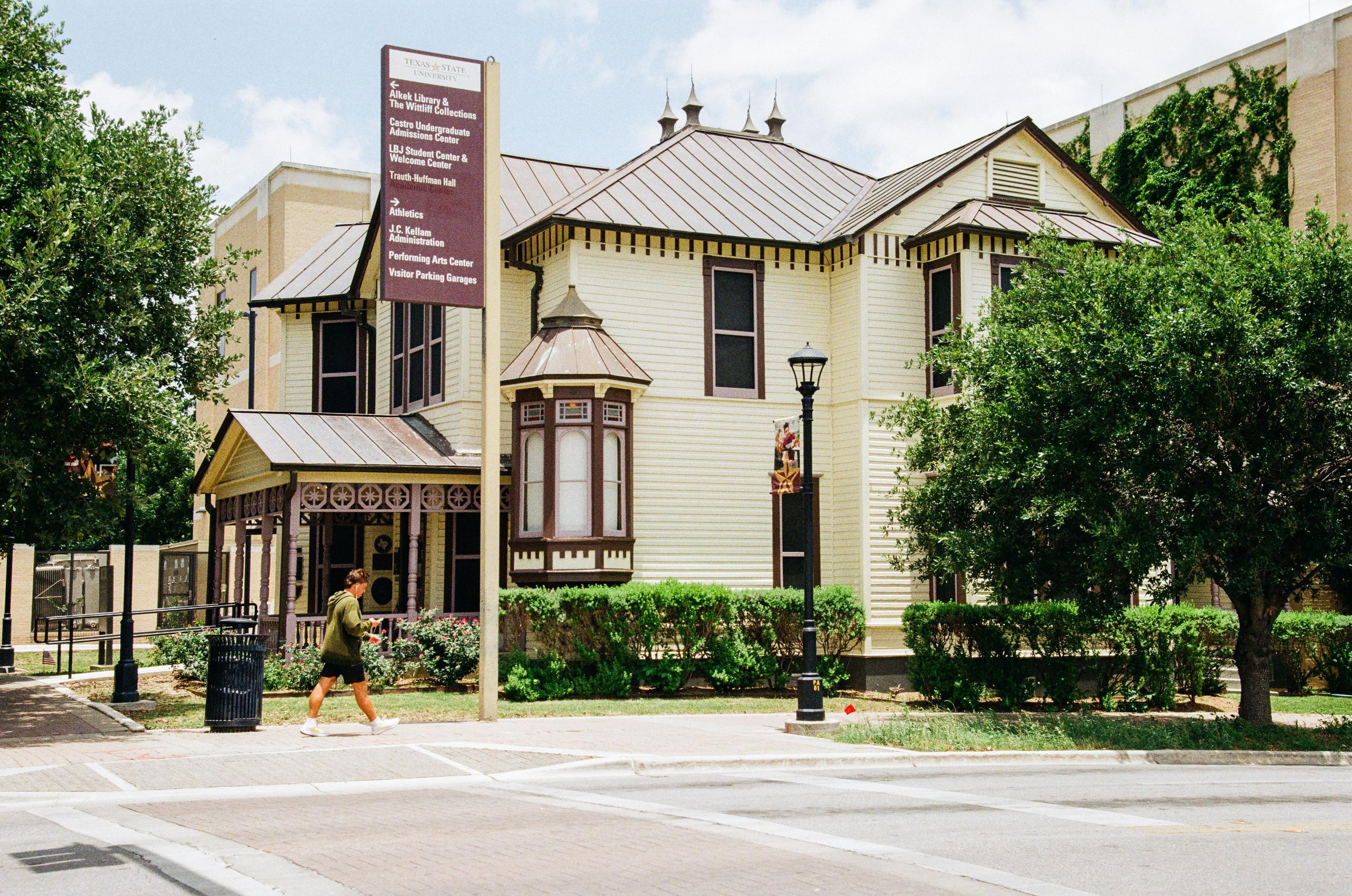 a man walks on the sidewalk in front of a pale yellow and maroon victorian-style house. 