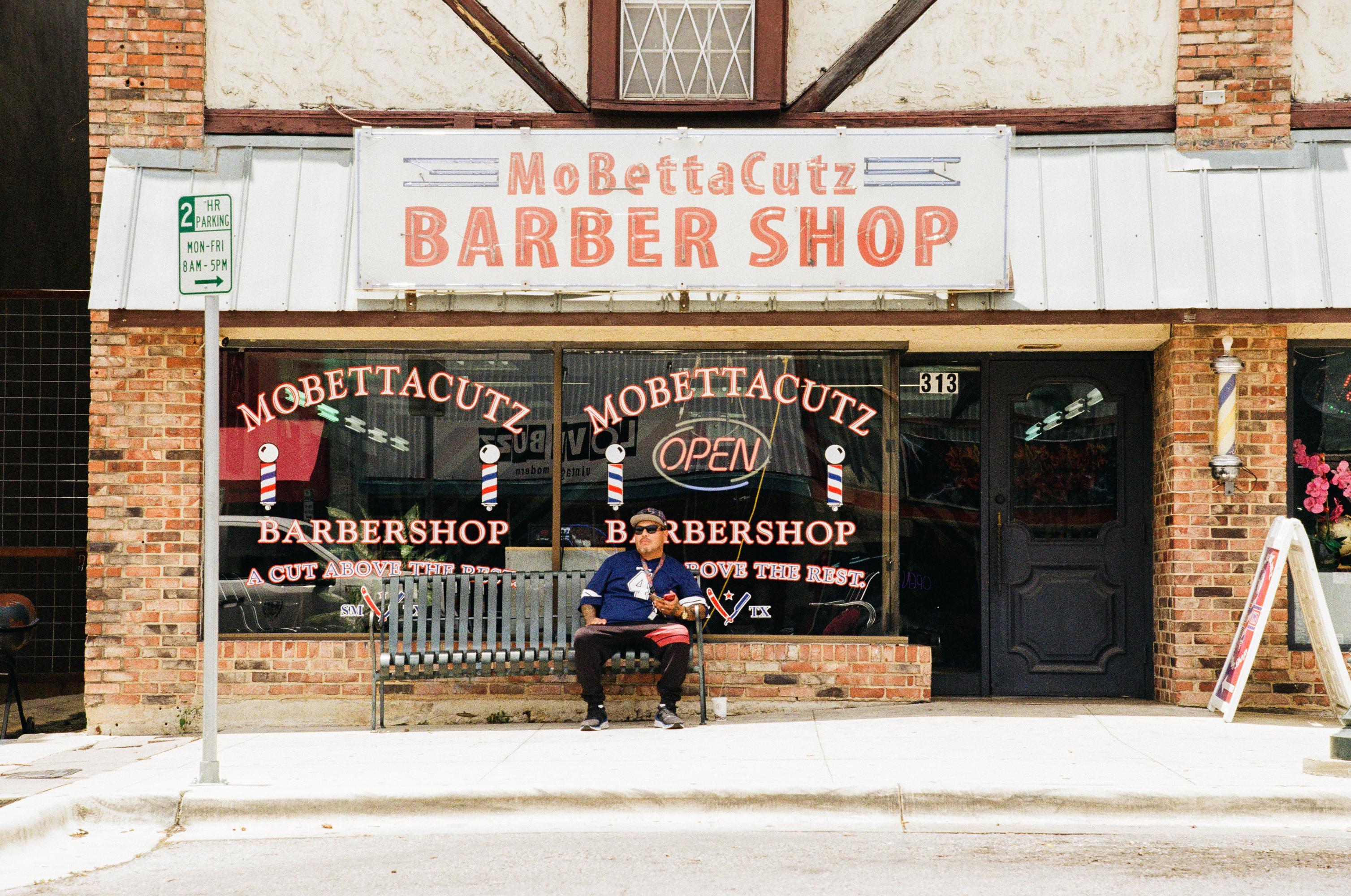 man in jeans and a dallas cowboys jersey sits on a bench in front of a barber shop. 