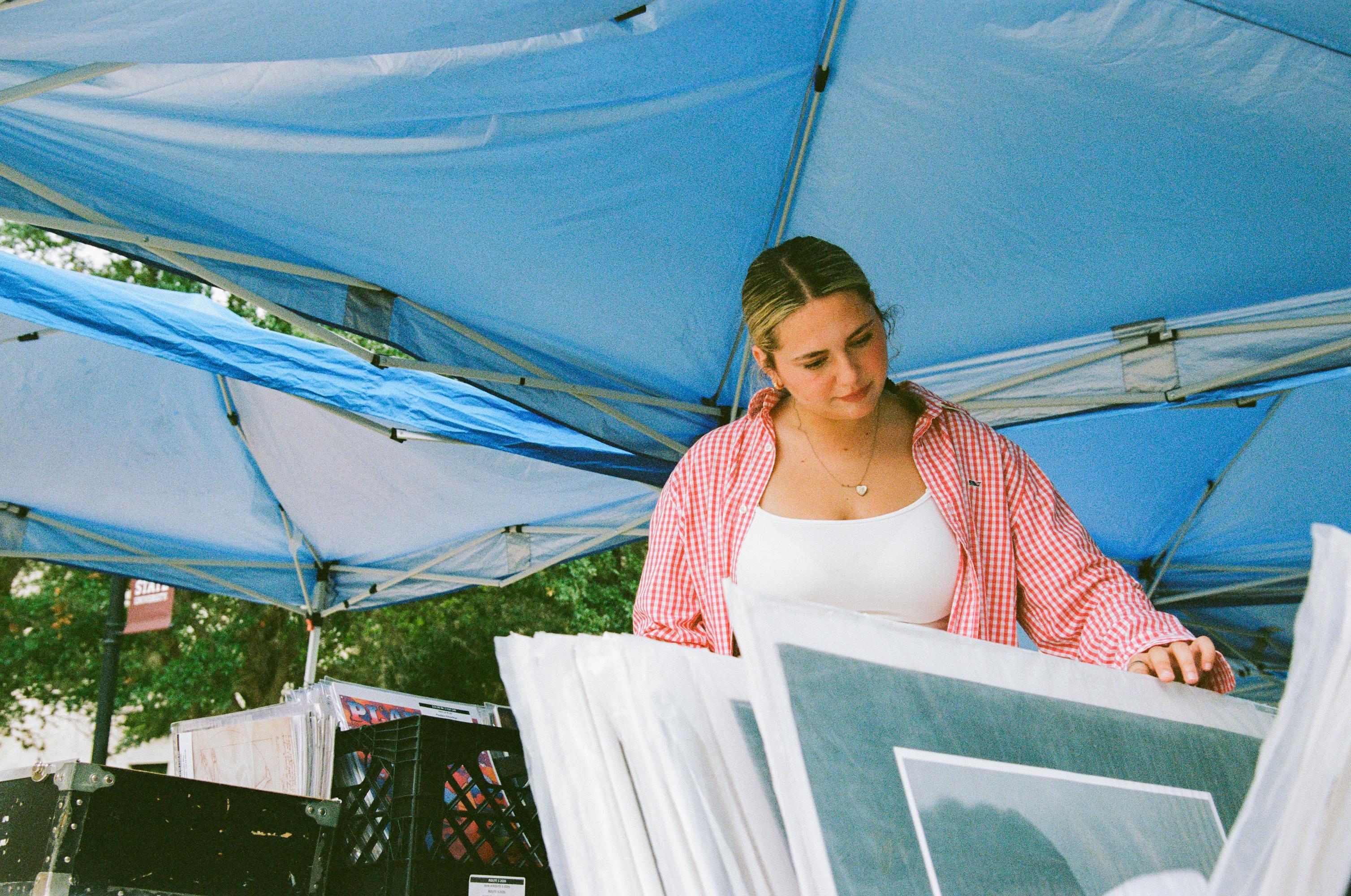 woman in white tank top and red gingham button down looks through set of art prints under a blue pop up tent 