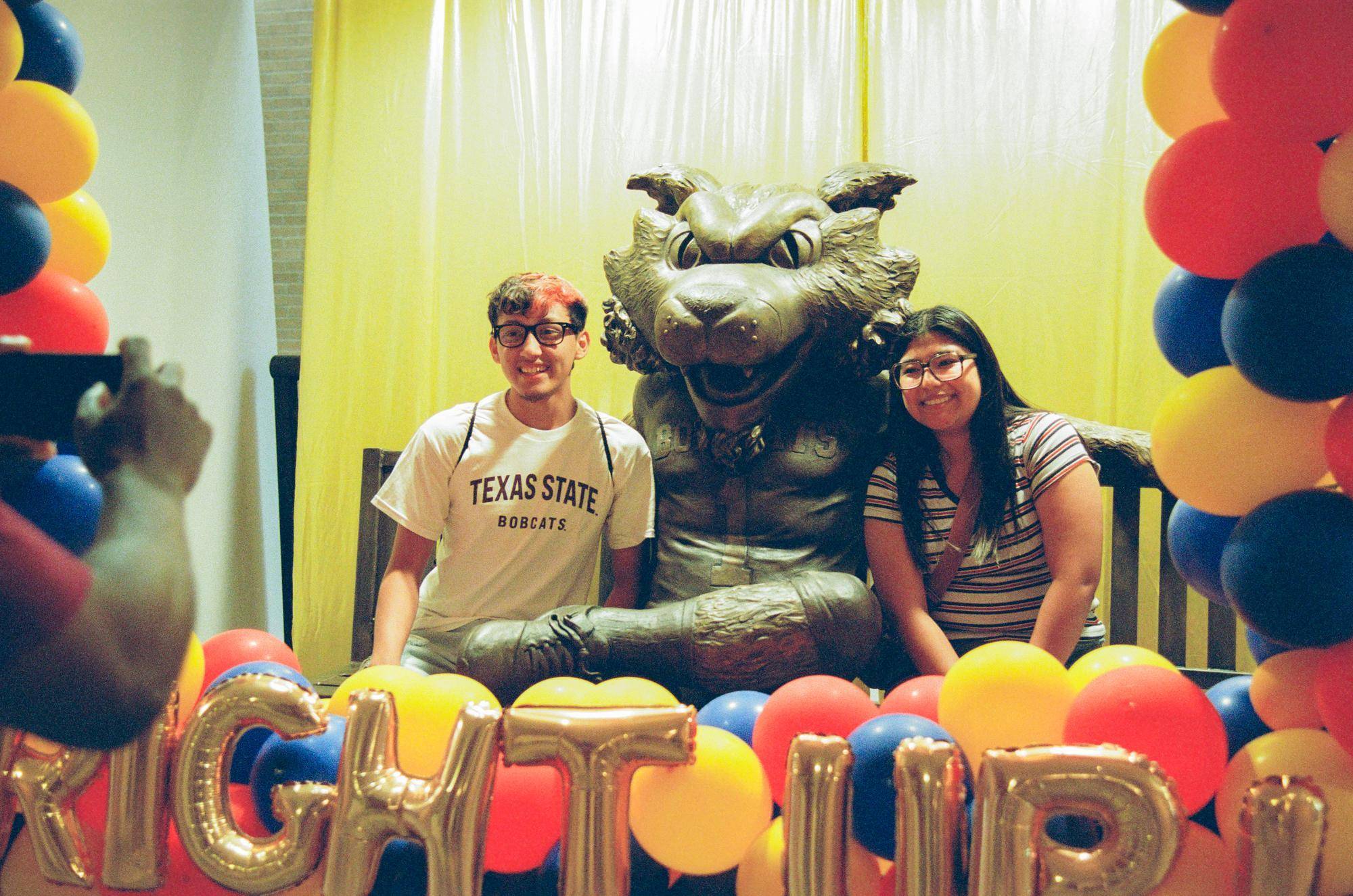 two students sit on a bench that features a large bronze statue of boko sitting down. there are balloon columns on either side of them