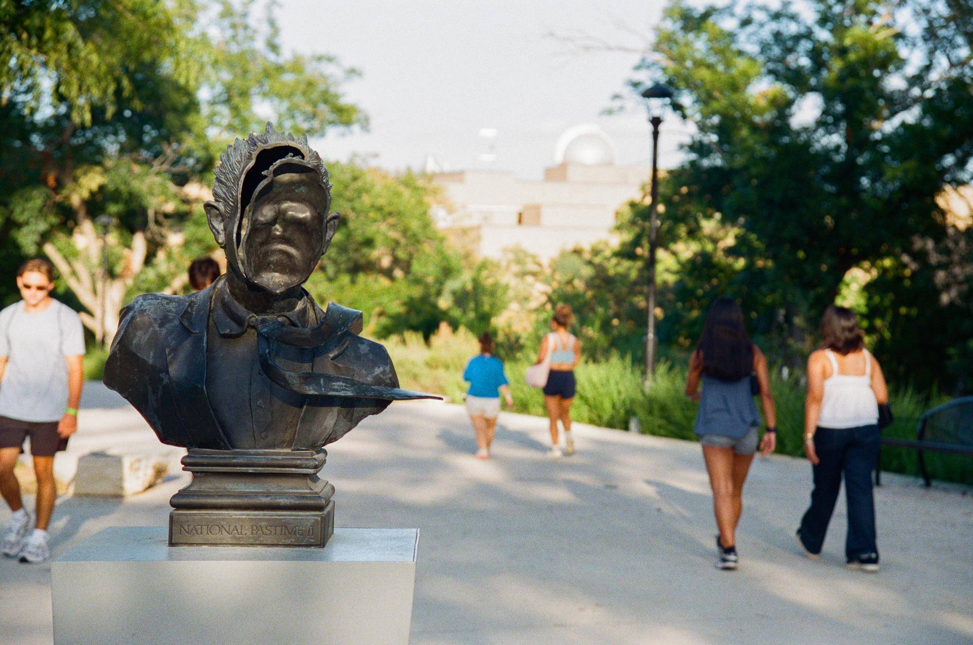 students walk on either side of a bronze statue. the statue is of a man's bust where the face is looking into its head