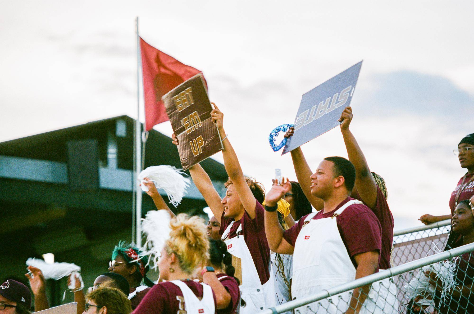multiple people in maroon shirts and white overalls hold up signs and cheer during a soccer game