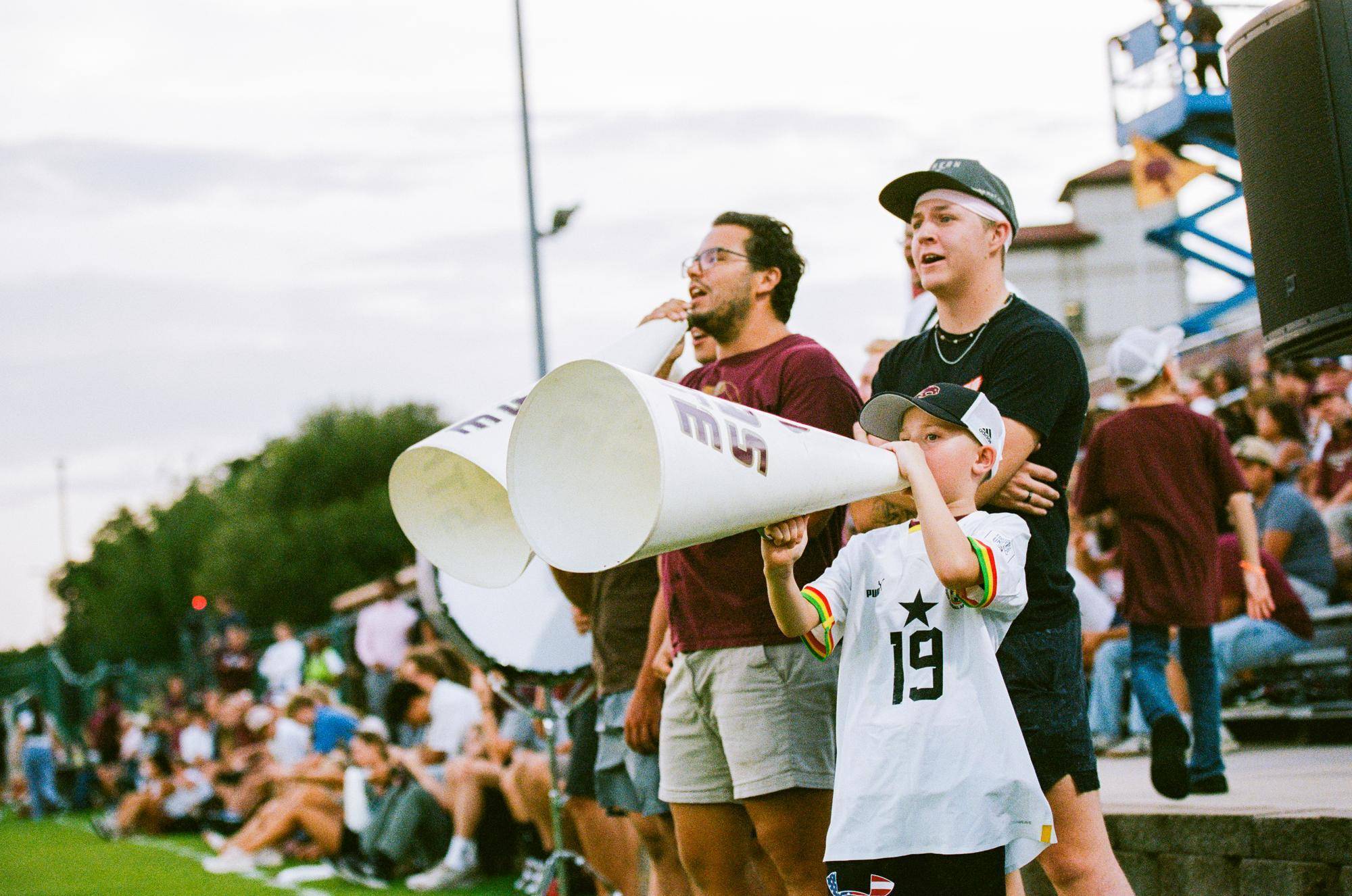 young boy is yelling into a cheerleader's megaphone while standing next to men cheering for a soccer game
