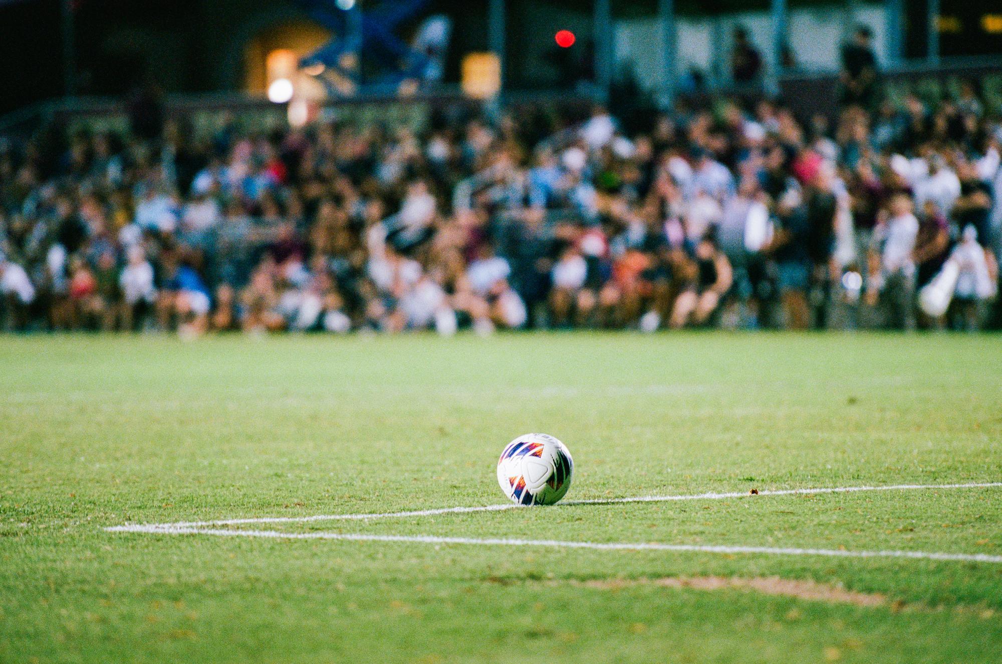 a soccer ball sits on an empty soccer field. a crowd of people watch in the distance. 