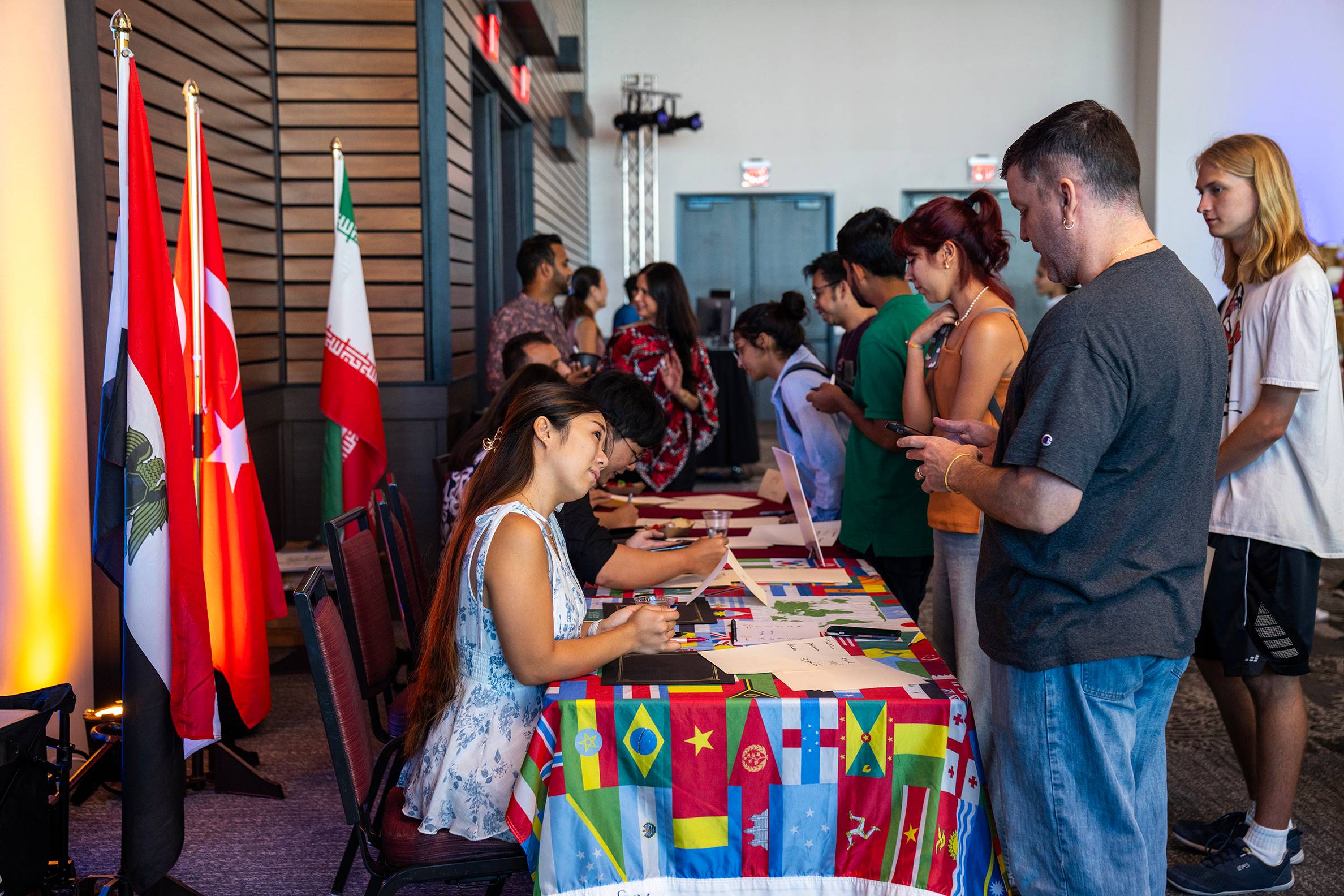 students at a tabling event for study abroad. international flags are lined up on a wall behind people sitting at tables talking to interested visitors. 