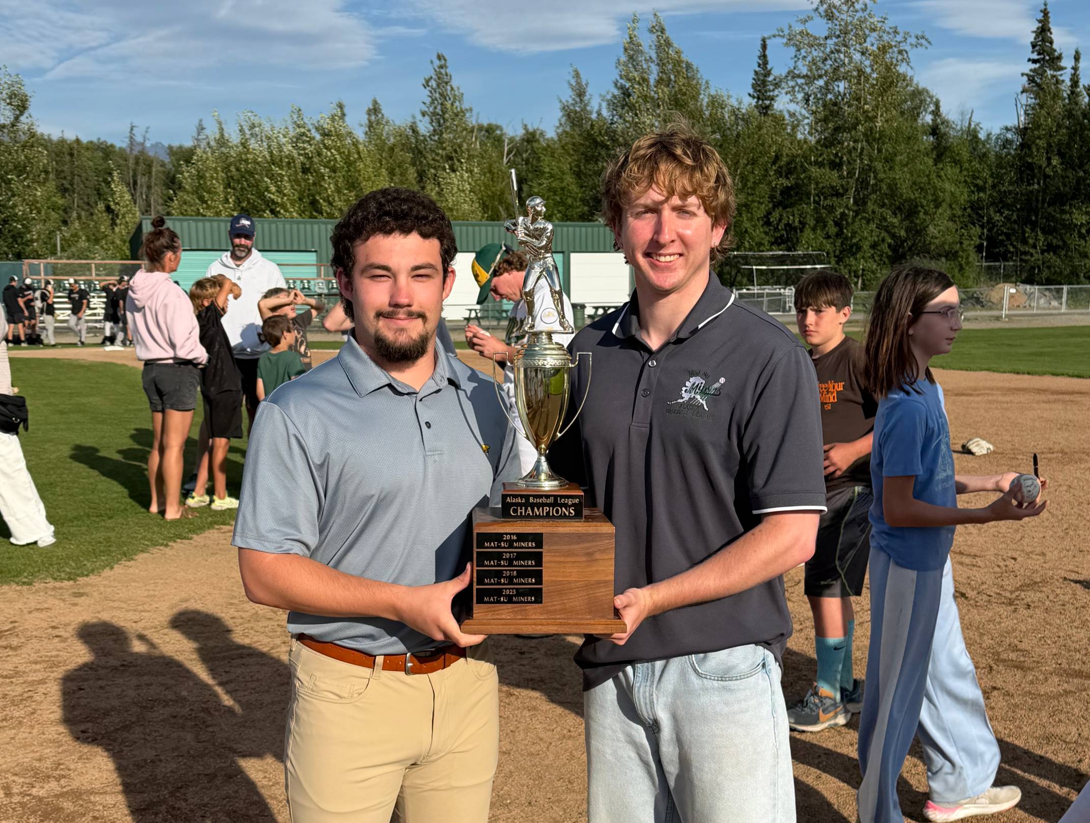two men in polo shirts hold a championship trophy while standing on a baseball field