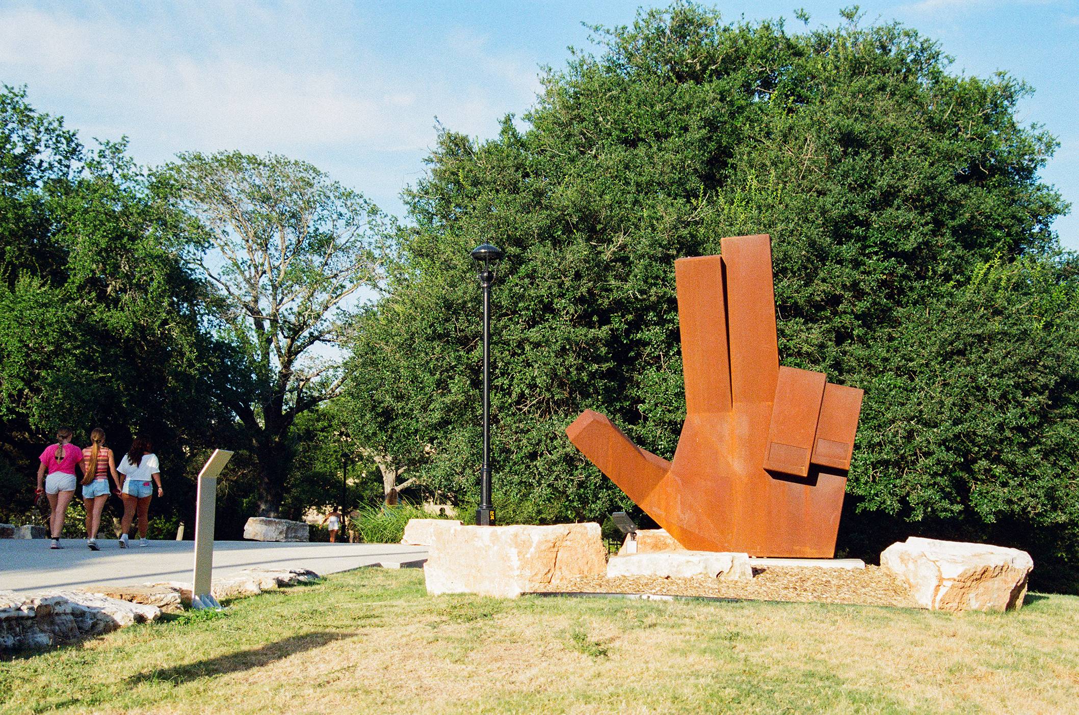 students walking on sidewalk next to large wooden sculpture of the txst hand sign
