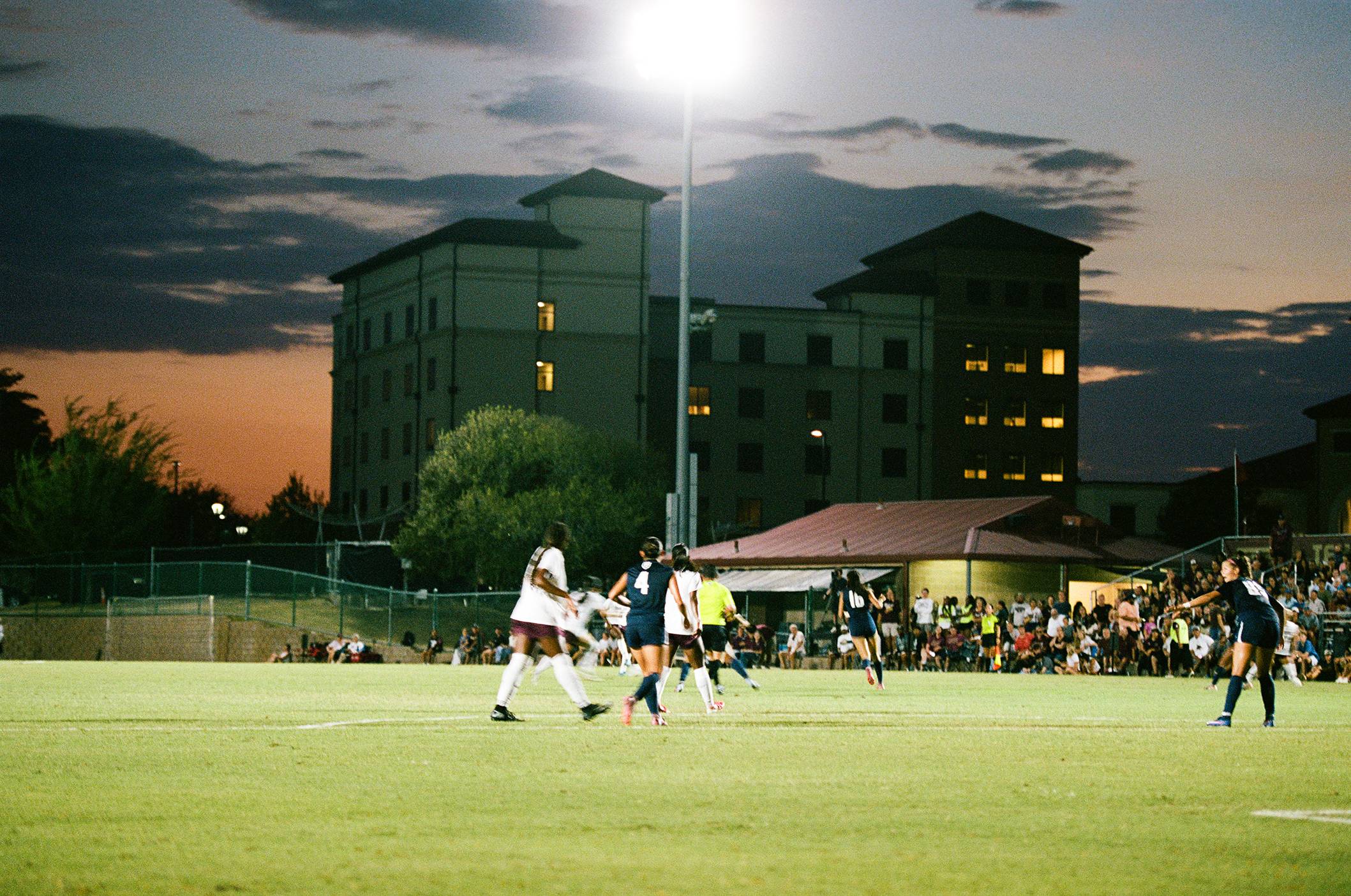 sun sets behind large building. a bright soccer game in happening in the foreground