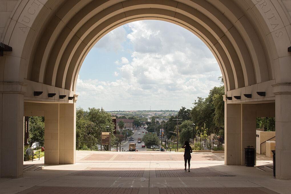 A view through a large stone archway overlooking a campus street, trees, buildings, and the surrounding landscape under a partly cloudy sky.