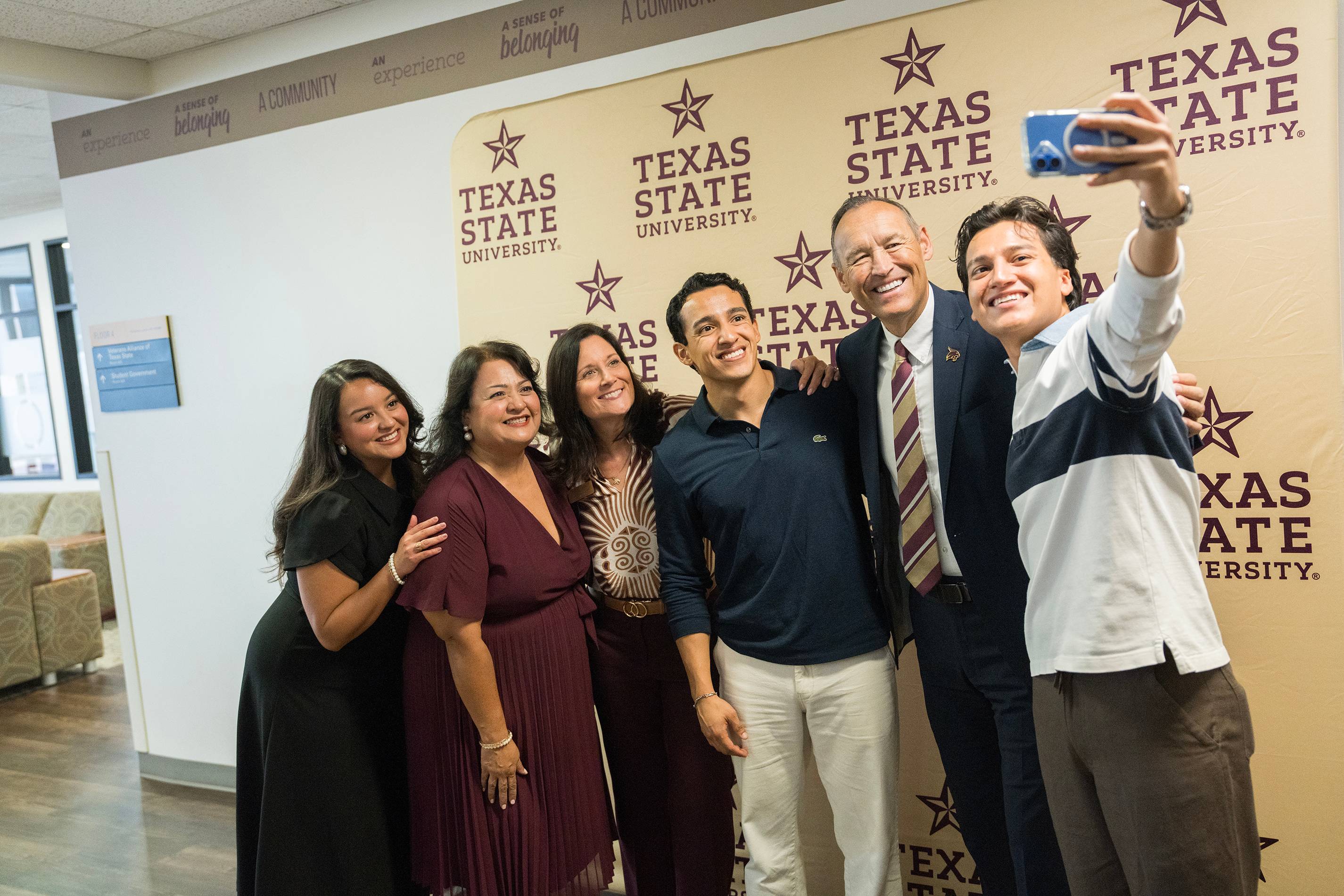 man takes selfie with five other individuals in front of gold texas state university banner