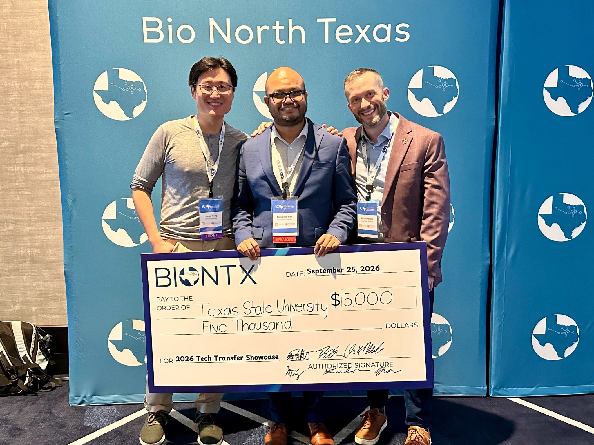 three men holding large check in front of blue wall reading "bio north texas"