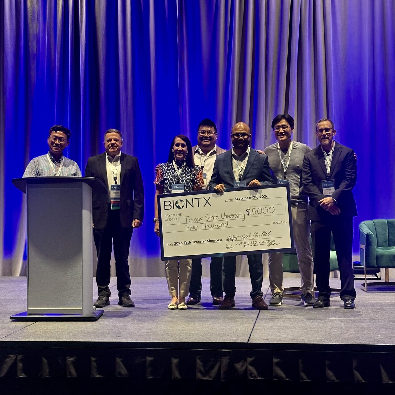 Dr. Aniruddha Bora and six others on stage holding an oversized $5,000 check from BIONTX made out to Texas State University for the 2026 Tech Transfer Showcase.