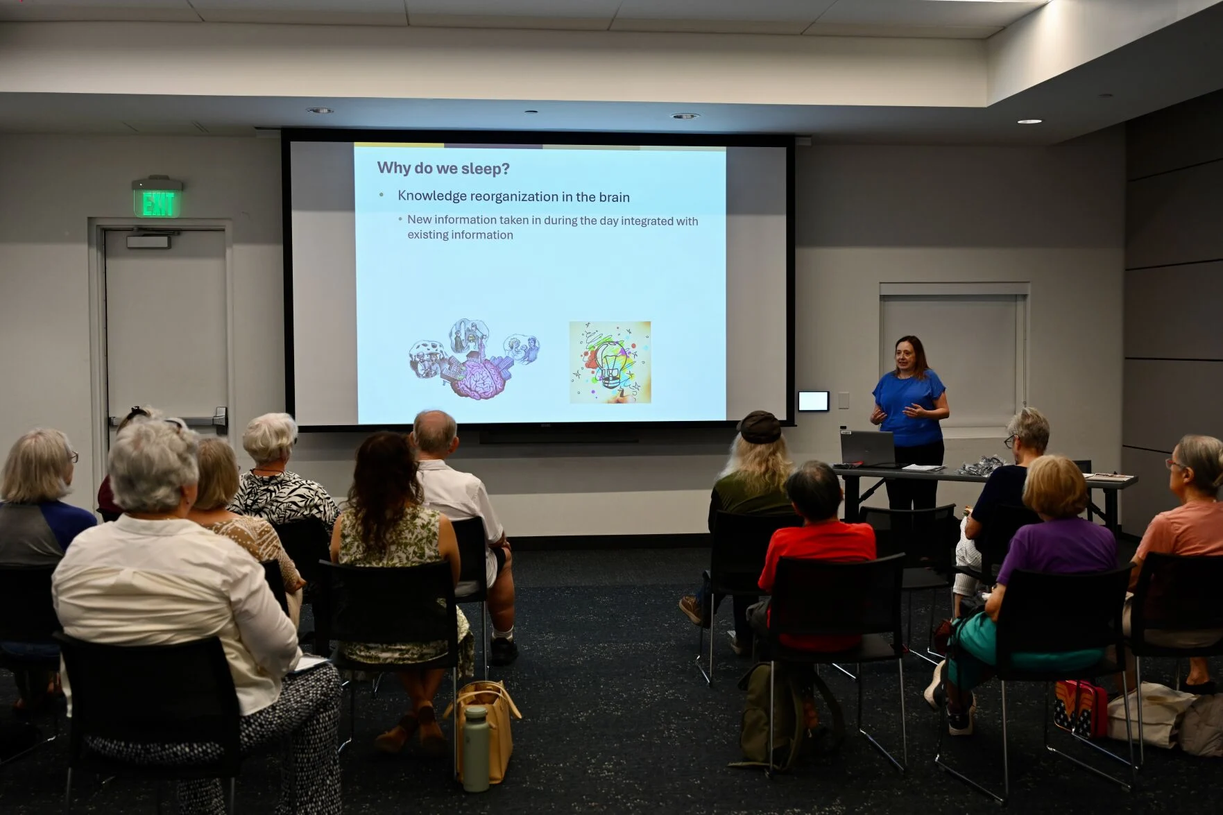 A presenter in a blue shirt addresses an audience seated in rows in a meeting room. Beside her, a projected slide titled “Why do we sleep?” explains knowledge reorganization in the brain, noting that new information taken in during the day is integrated with existing information. Illustrations of a brain and a colorful light bulb appear beneath the text. A laptop and presentation materials sit on a table in front of her.