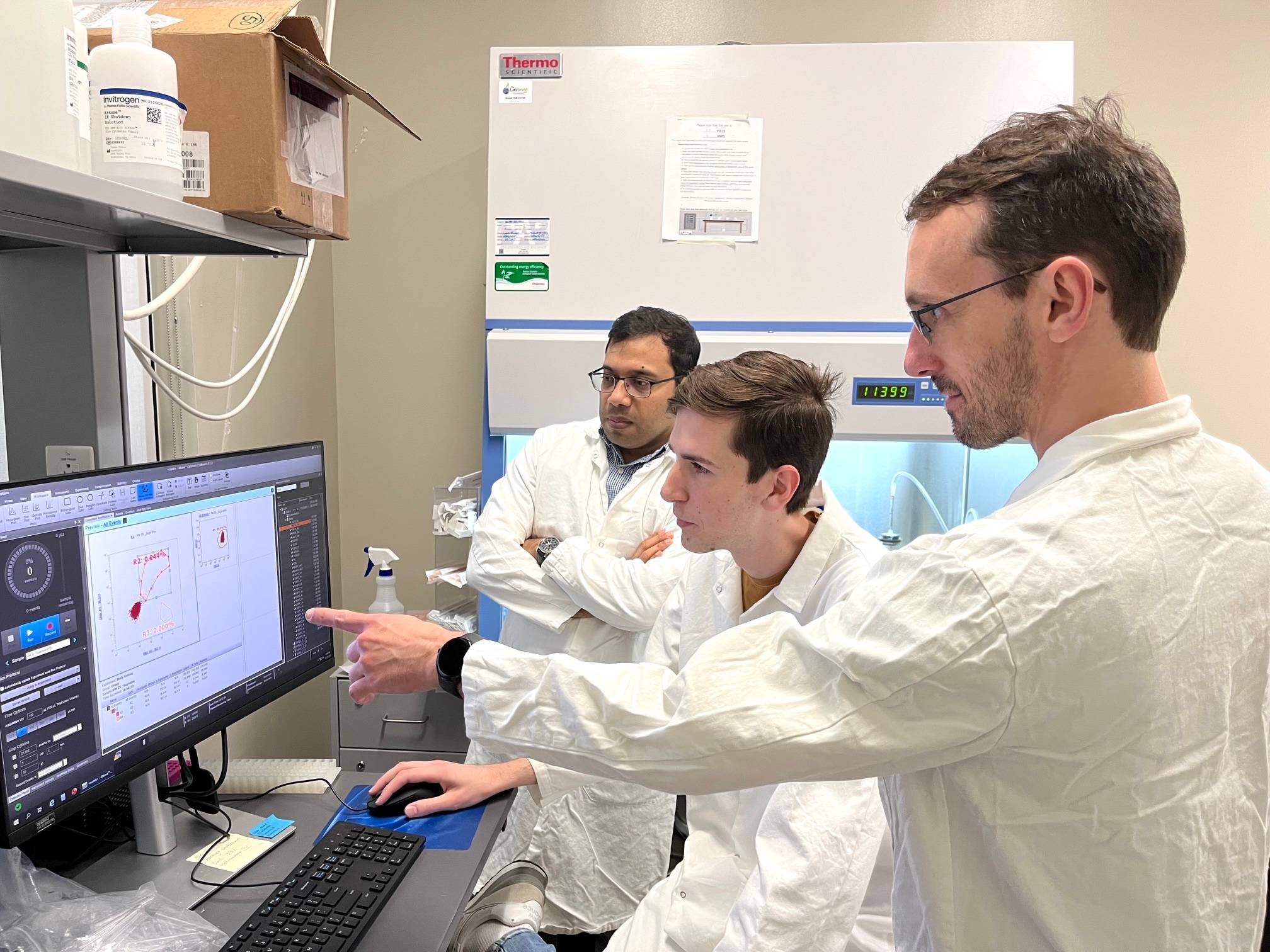 three men in white lab coats look at computer monitor