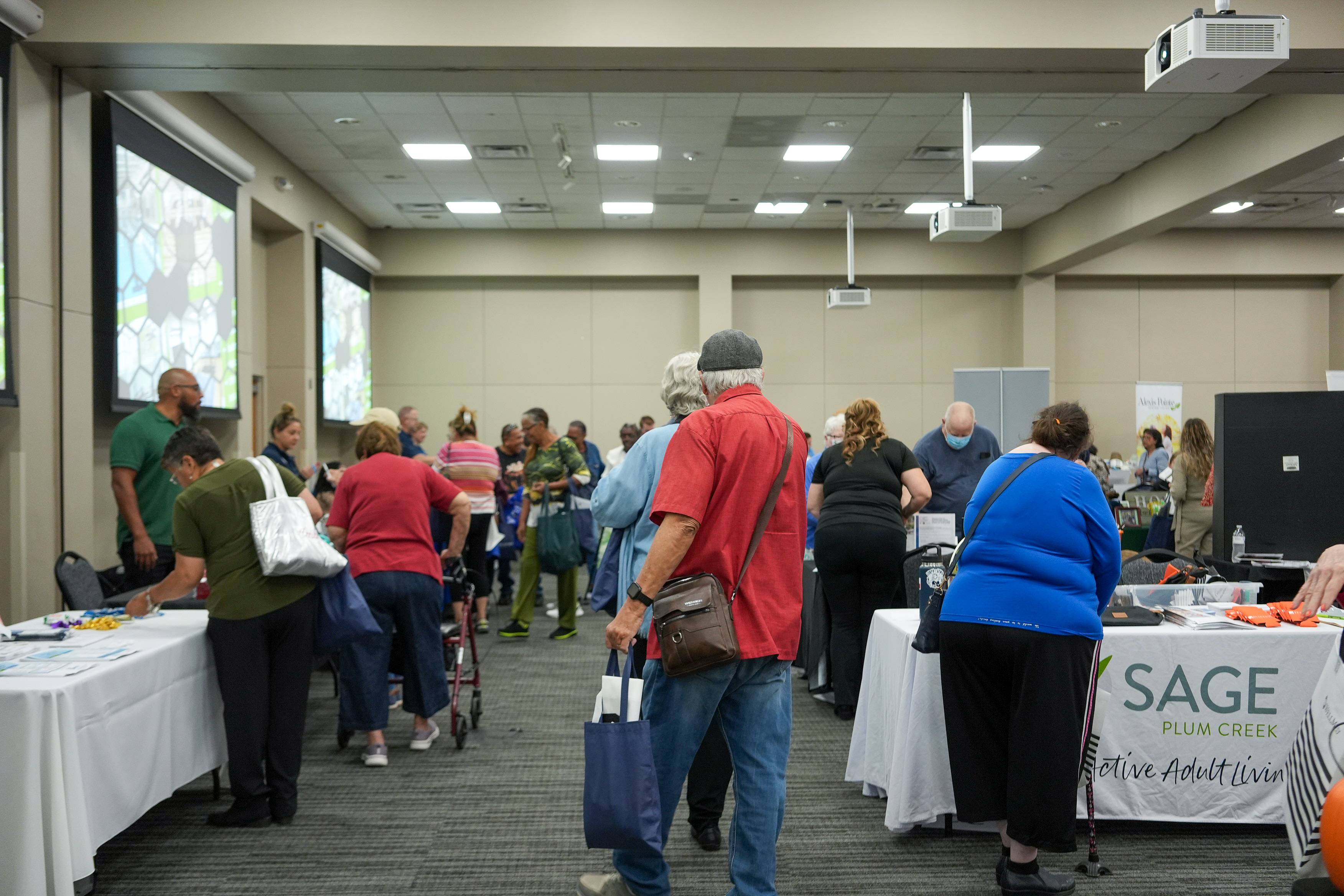 A busy room at the 12th Senior Citizen Expo at the San Marcos Activity Center. Older adults, some carrying tote bags and one using a walker, move between rows of exhibitor tables covered in brochures and giveaways while vendors talk with visitors.