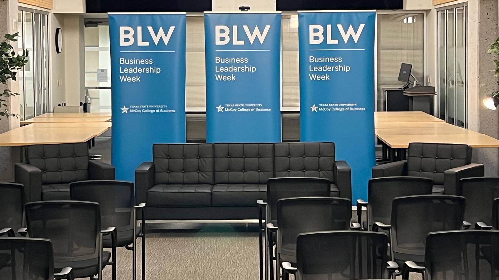 An event space at Texas State University's McCoy College of Business set up for Business Leadership Week, featuring rows of chairs facing a seating area with a black couch and armchairs, backed by three blue Business Leadership Week banners.